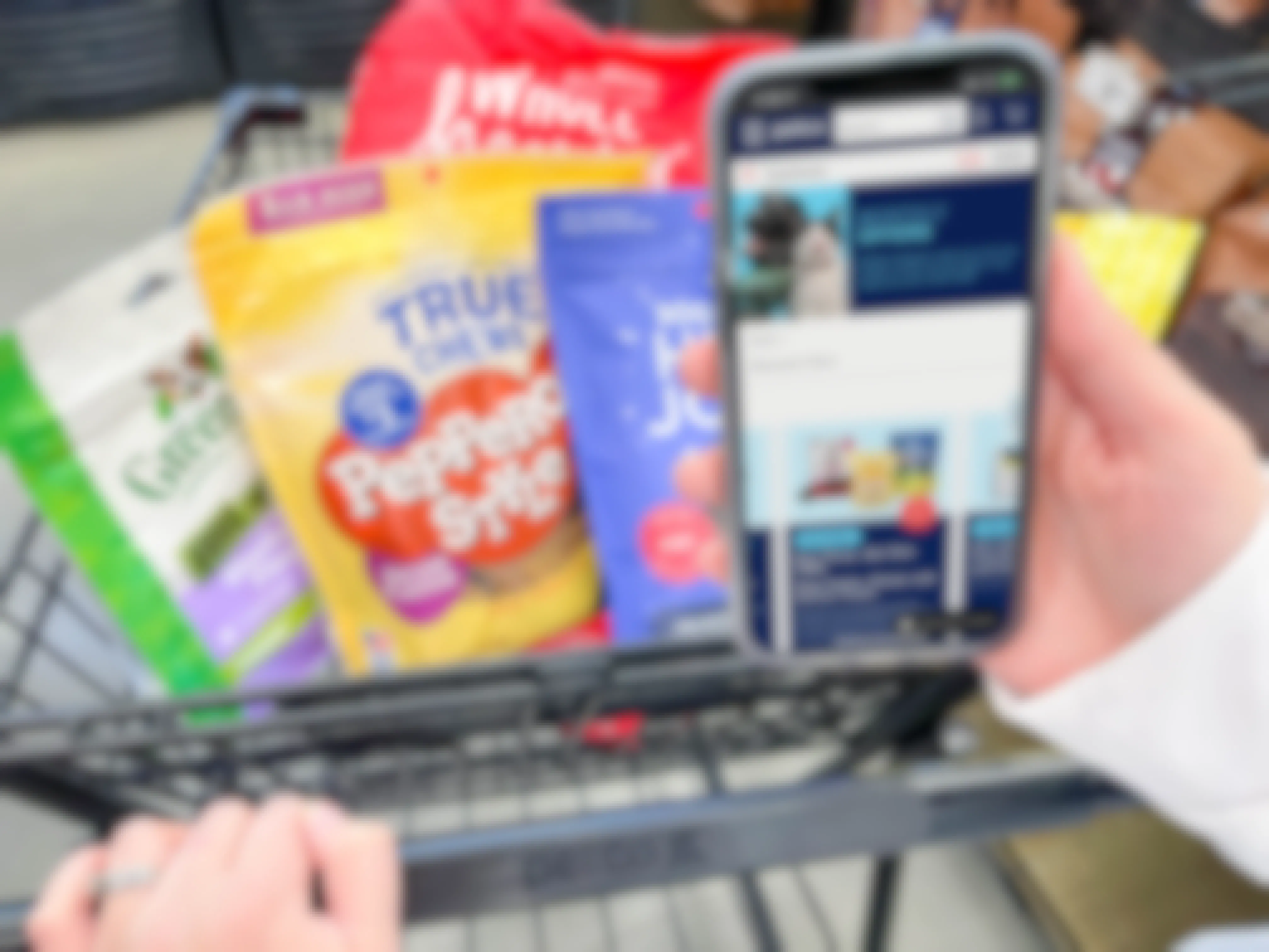 a person holding a cellphone with petco monthly coupons on screen while pushing cart with dog treats 