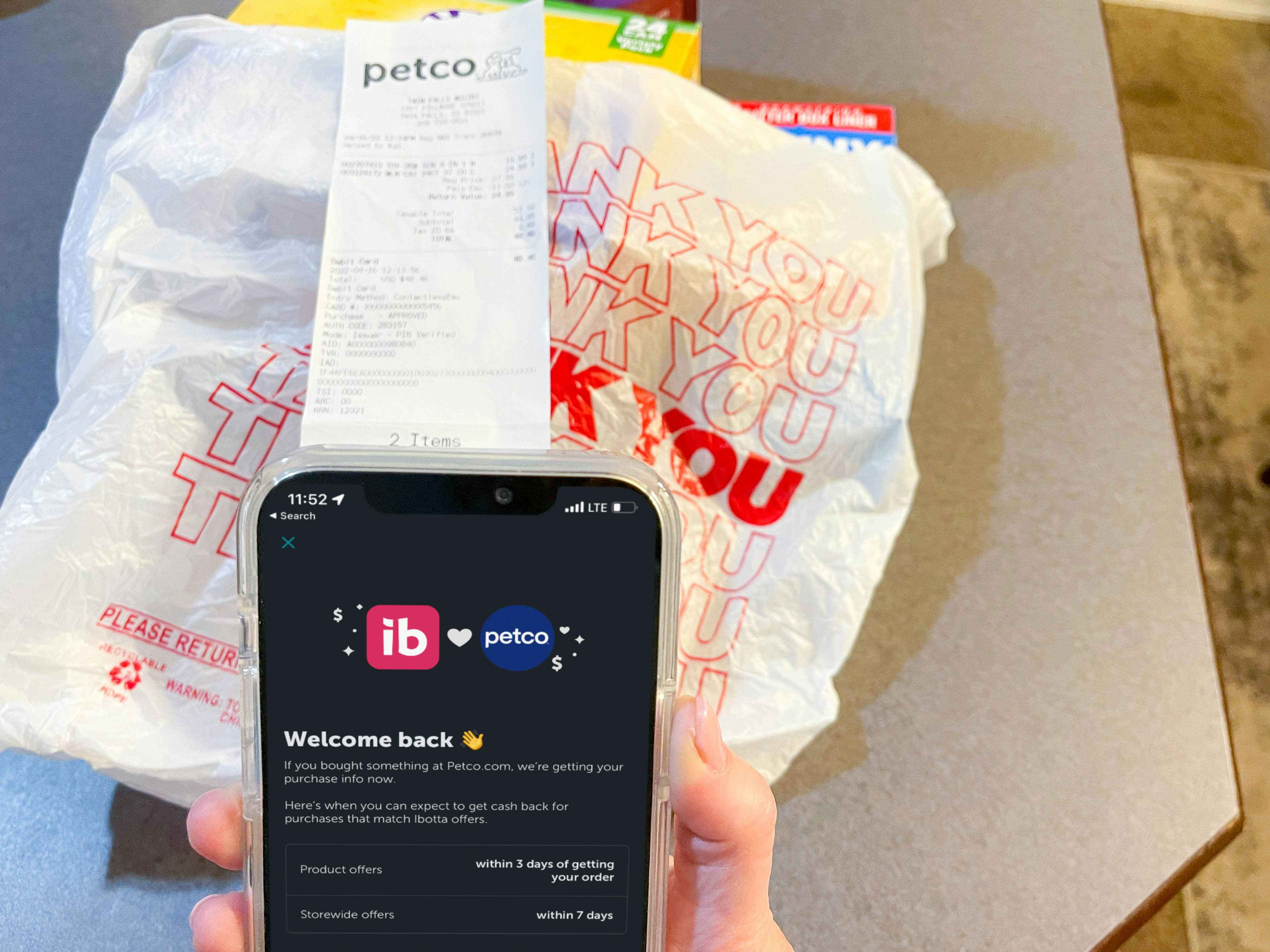 a person holding a cellphone with ibotta app ad petco on screen with bag of items and petco receipt in background
