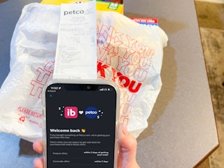 a person holding a cellphone with ibotta app ad petco on screen with bag of items and petco receipt in background