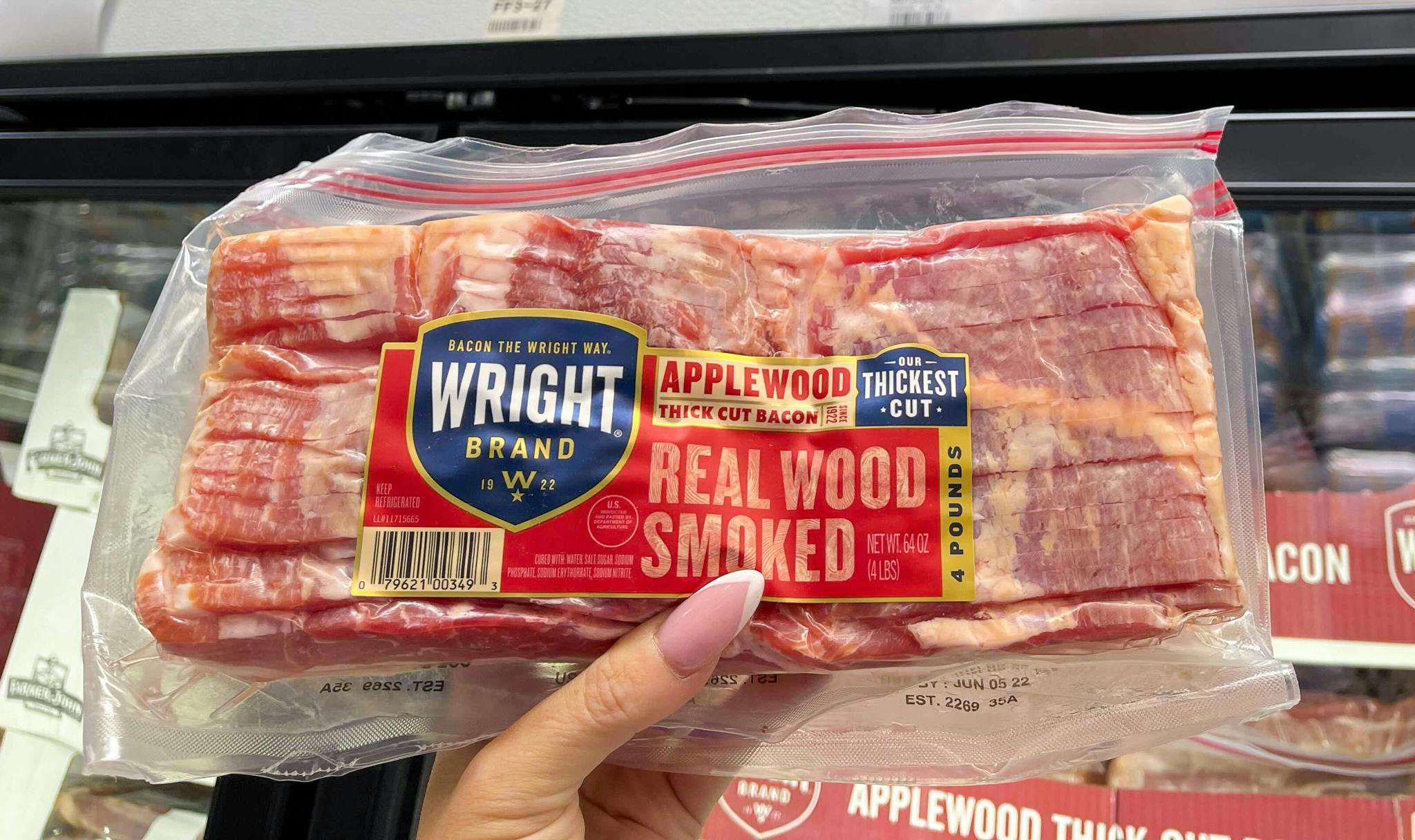 A person's hand holding up a package of Wright Brand real wood smoked bacon in the refrigerated section at Sam's Club.