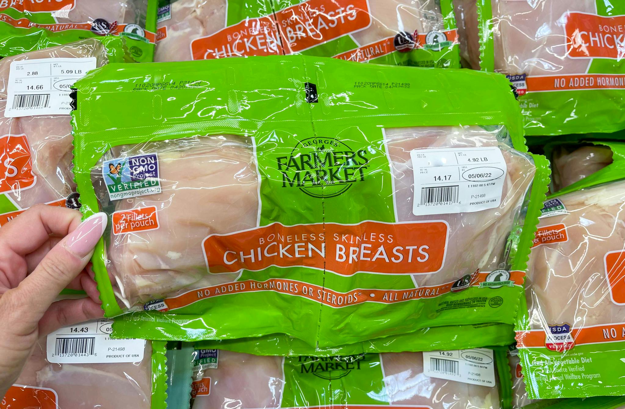 A person's hand lifting a two-pack of Farmer's Market chicken breasts off of a shelf of more of the same product at Sam's Club.