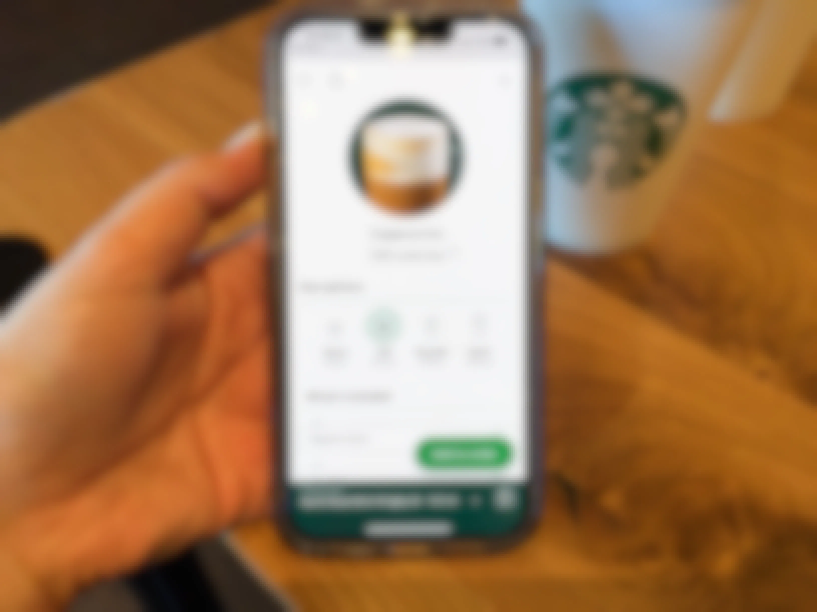 A person's hand holding an iPhone displaying the Starbucks app's page for ordering a Cappuccino next to a Starbucks drink cup on a table.