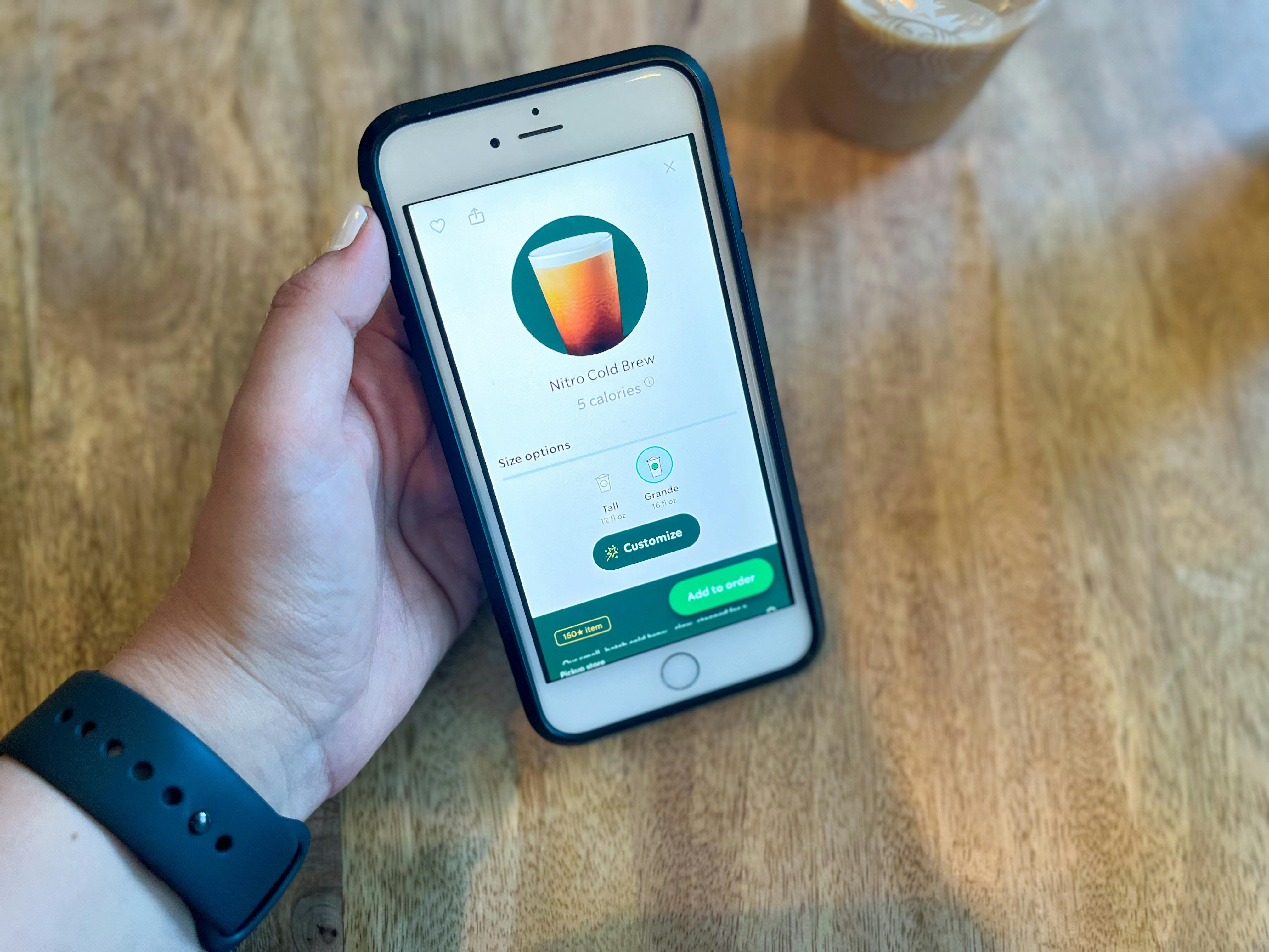 A person's hand holding an iPhone displaying the Starbucks app's page to order a Grande Nitro Cold Brew next to a Starbucks cup.