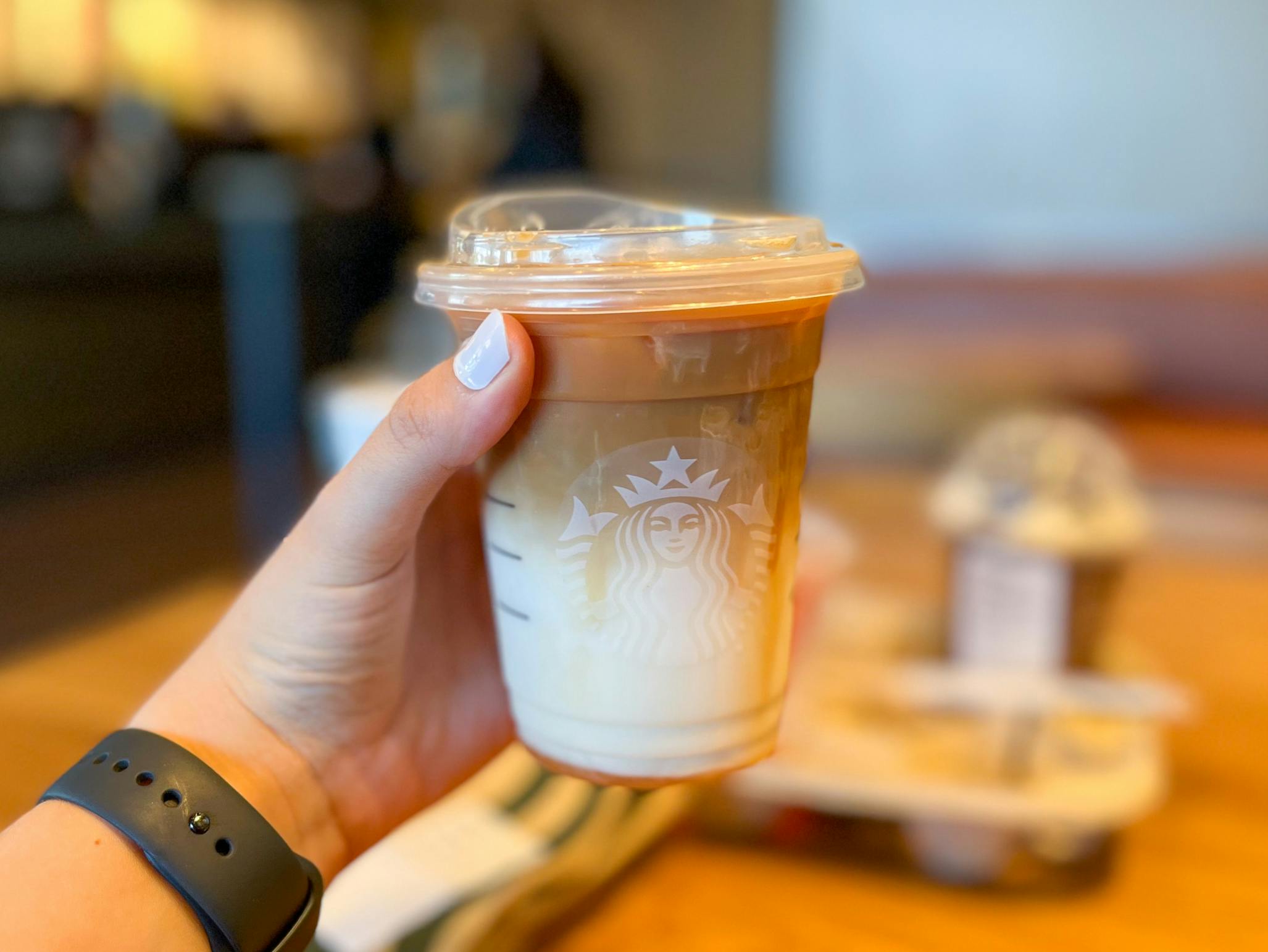 A person's hand holding up a Grande Caramel Macchiato in front of a table of more drinks at Starbucks.