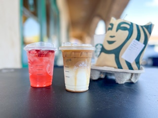 Two Starbucks Grande-sized drinks sitting next to a paper Starbucks bag on a table outside of Starbucks.