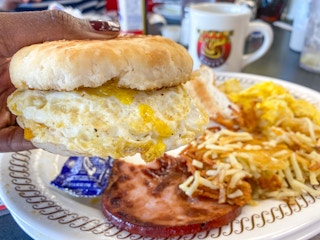 A person's hand holding up a sausage, egg, and cheese biscuit sandwich above a plate with hash browns at a Waffle House.