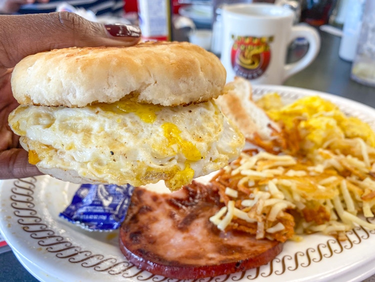 A person's hand holding up a sausage, egg, and cheese biscuit sandwich above a plate with hash browns at a Waffle House.