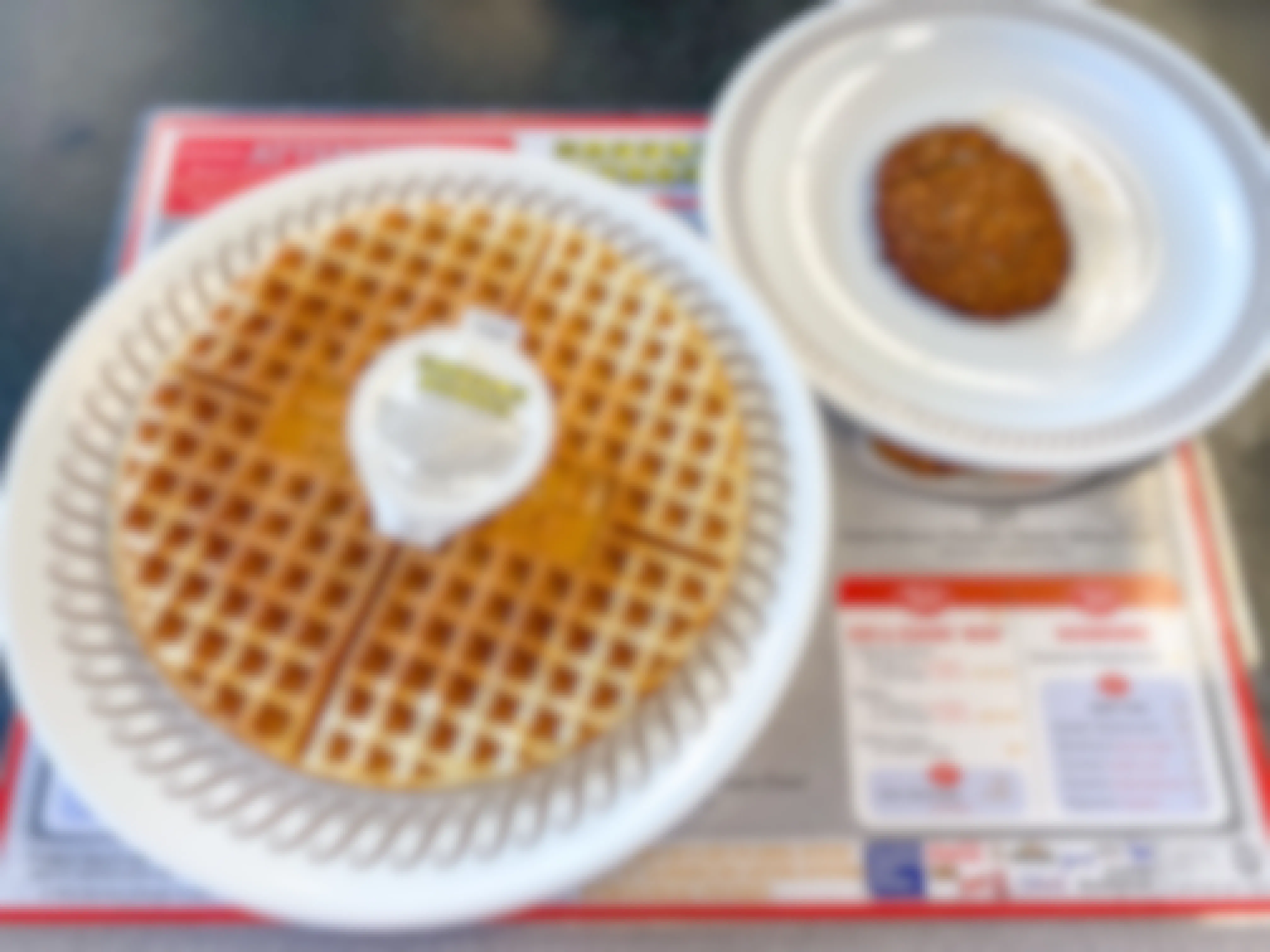 A plate with a big waffle and a container of Waffle House butter sitting next to another plate with a sausage patty on a table at Waffle House.