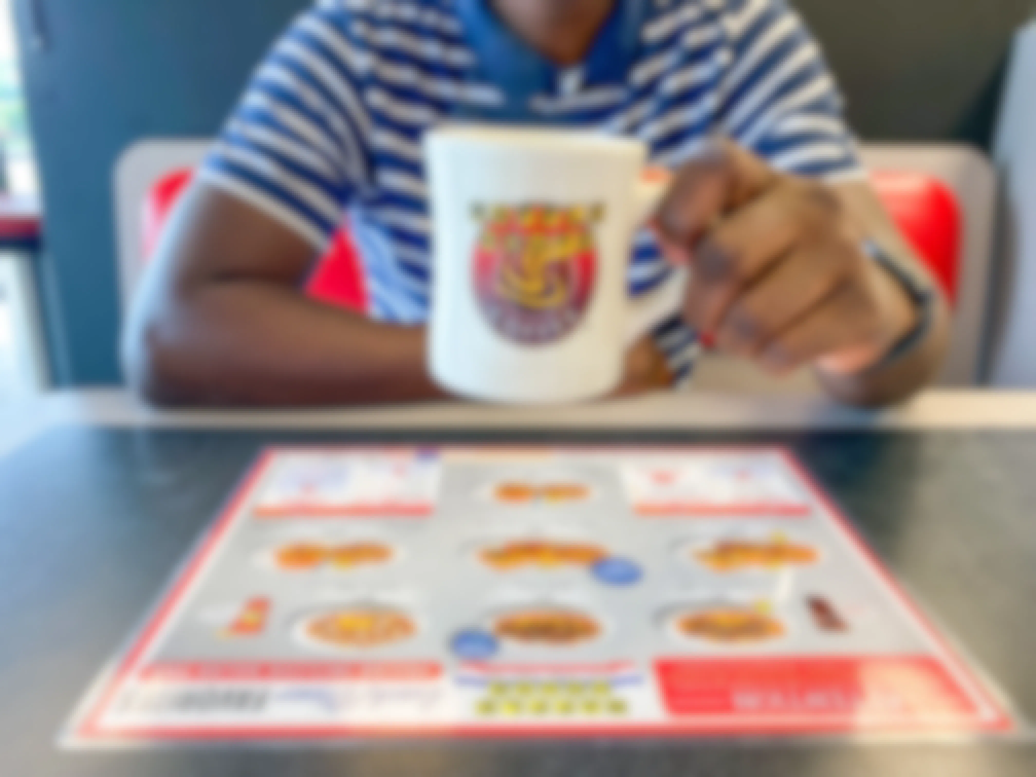 A person holding up a Waffle House coffee mug while sitting at a booth inside Waffle House.