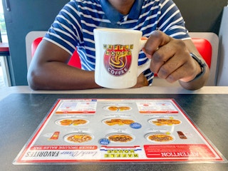 A person holding up a Waffle House coffee mug while sitting at a booth inside Waffle House.
