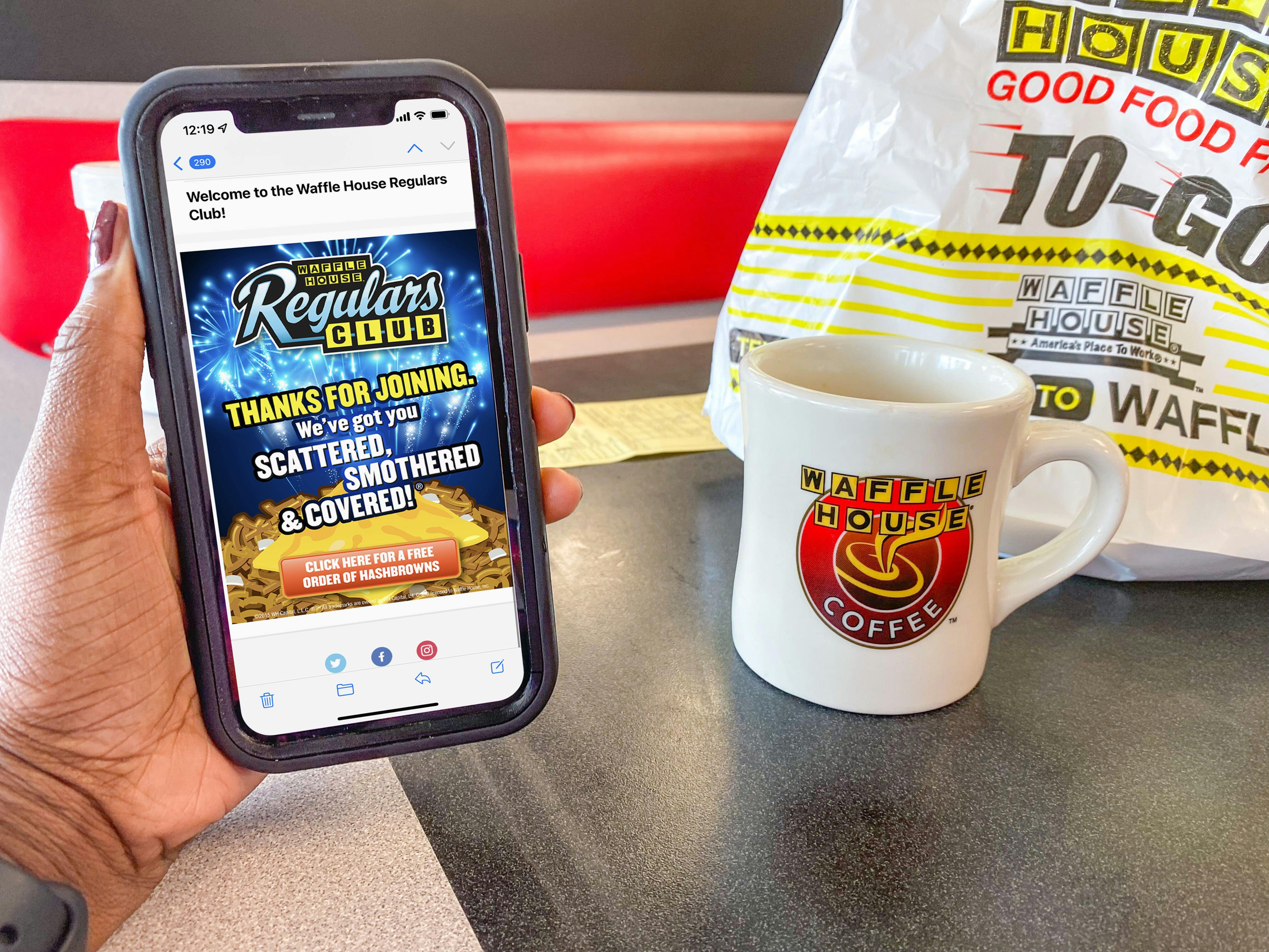 A person's hand holding a cell phone displaying a welcome email from Waffle House Regular Club in front of a Waffle House mug and to-go bag.