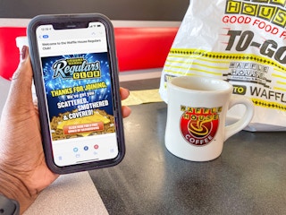 A person's hand holding a cell phone displaying a welcome email from Waffle House Regular Club in front of a Waffle House mug and to-go bag.