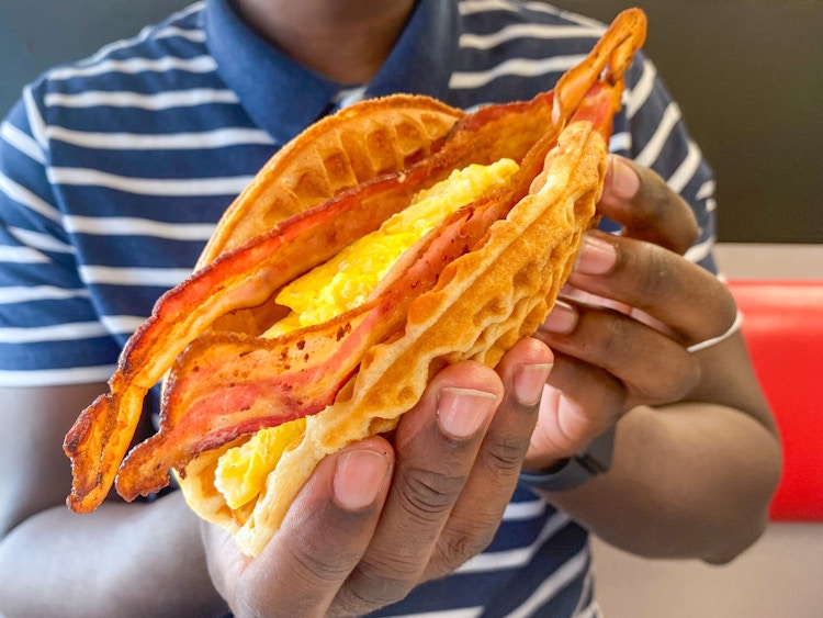 A person sitting in a booth at Waffle House, holding up a waffle sandwich with eggs and bacon.
