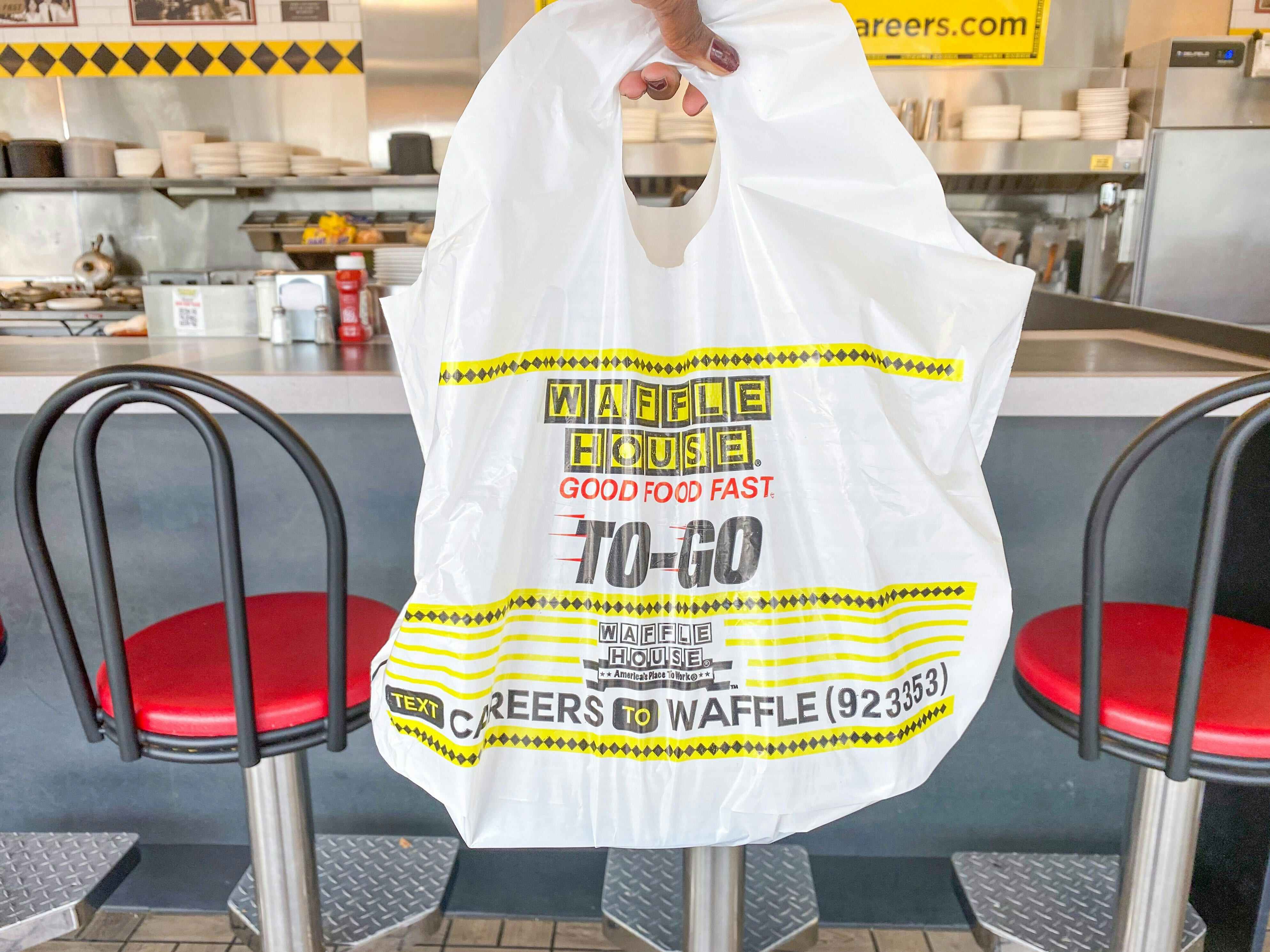 A person's hand holding up a Waffle House takeout bag in front of the counter at Waffle House.