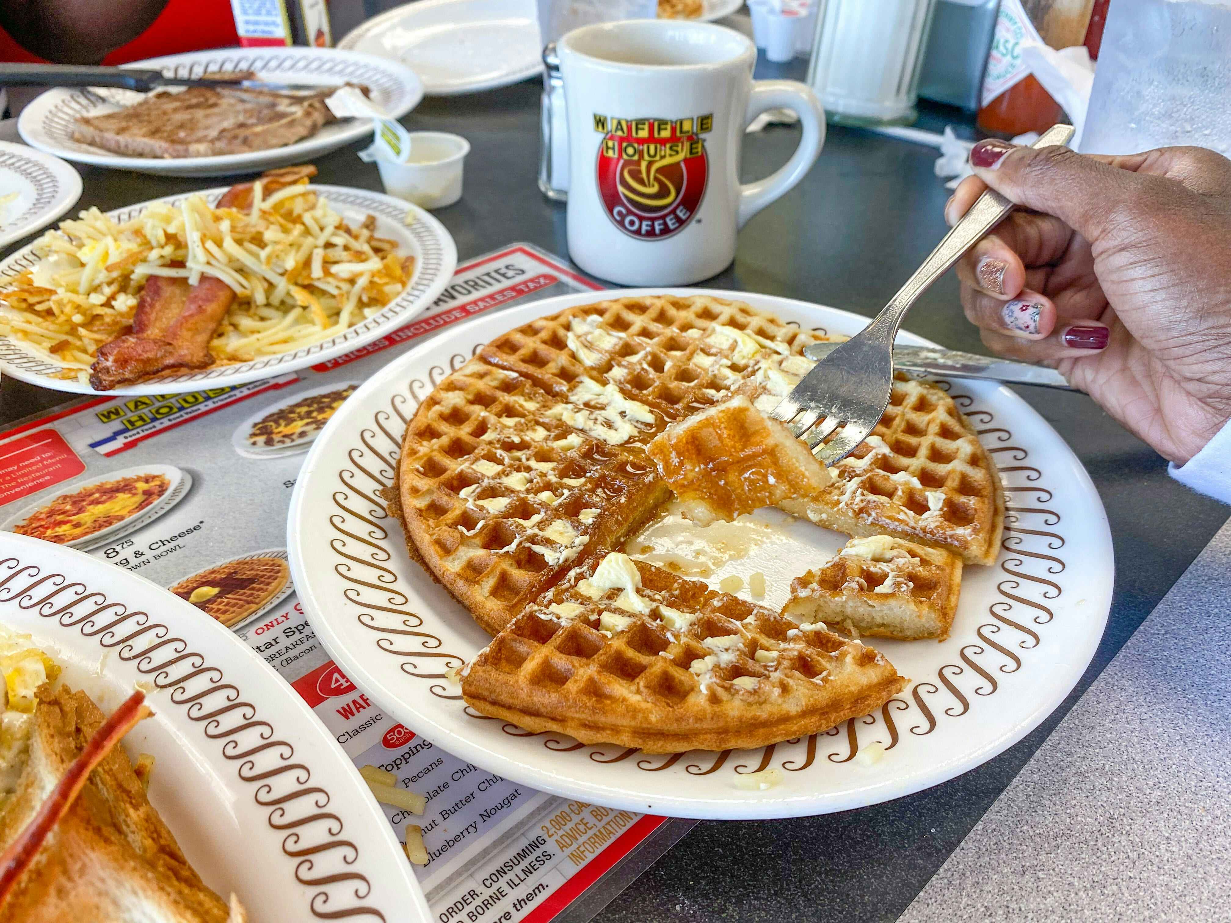 A person's hand using a fork to pick up a piece of a waffle from a plate on a Waffle House table next to other plated breakfast items.