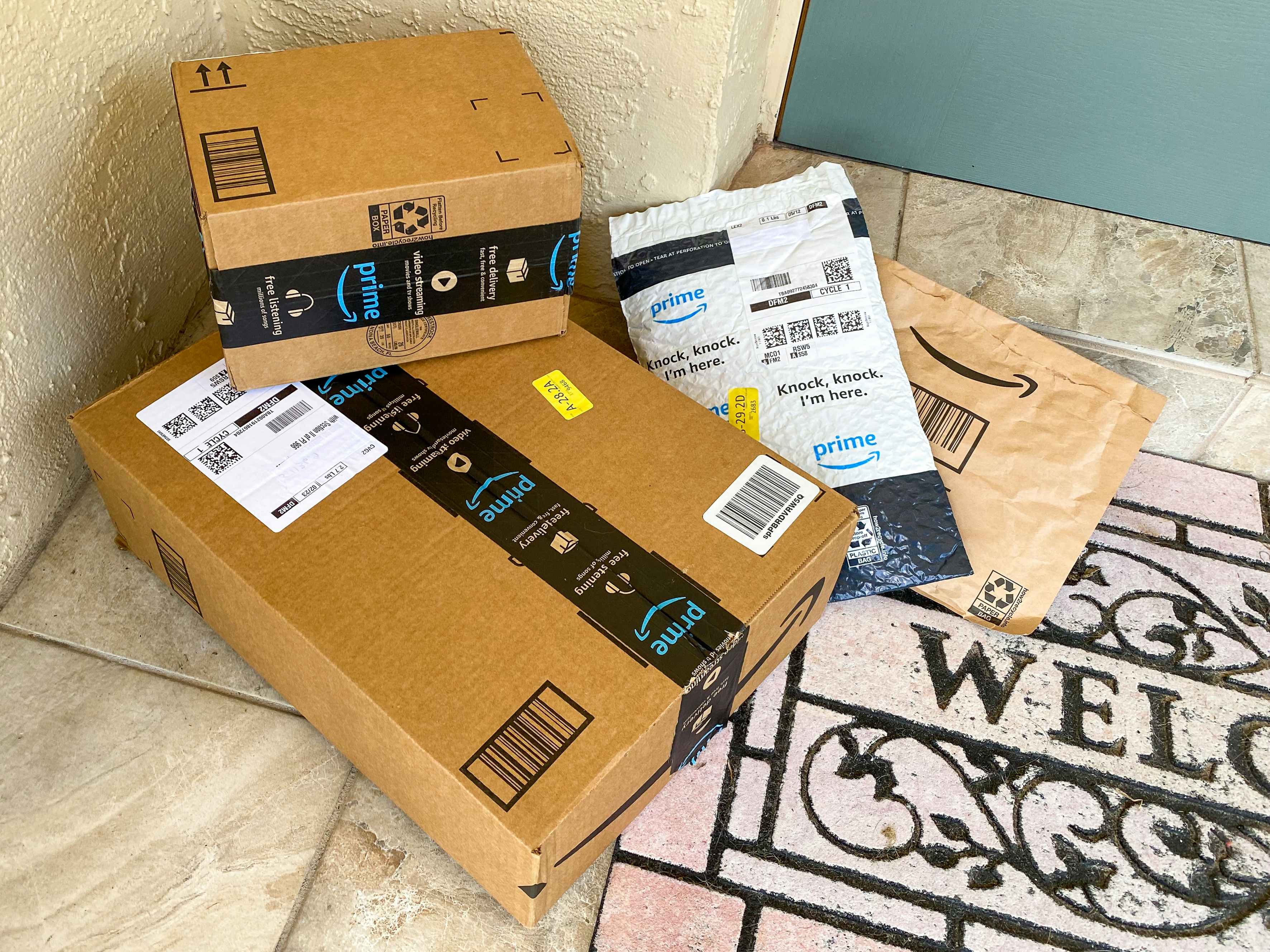 A pile of Amazon boxes and Amazon packing envelopes sitting on a doormat outside a front door.