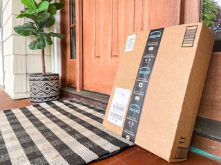 An Amazon box sitting on a front porch, leaning against the front door frame, next to a potted plant.