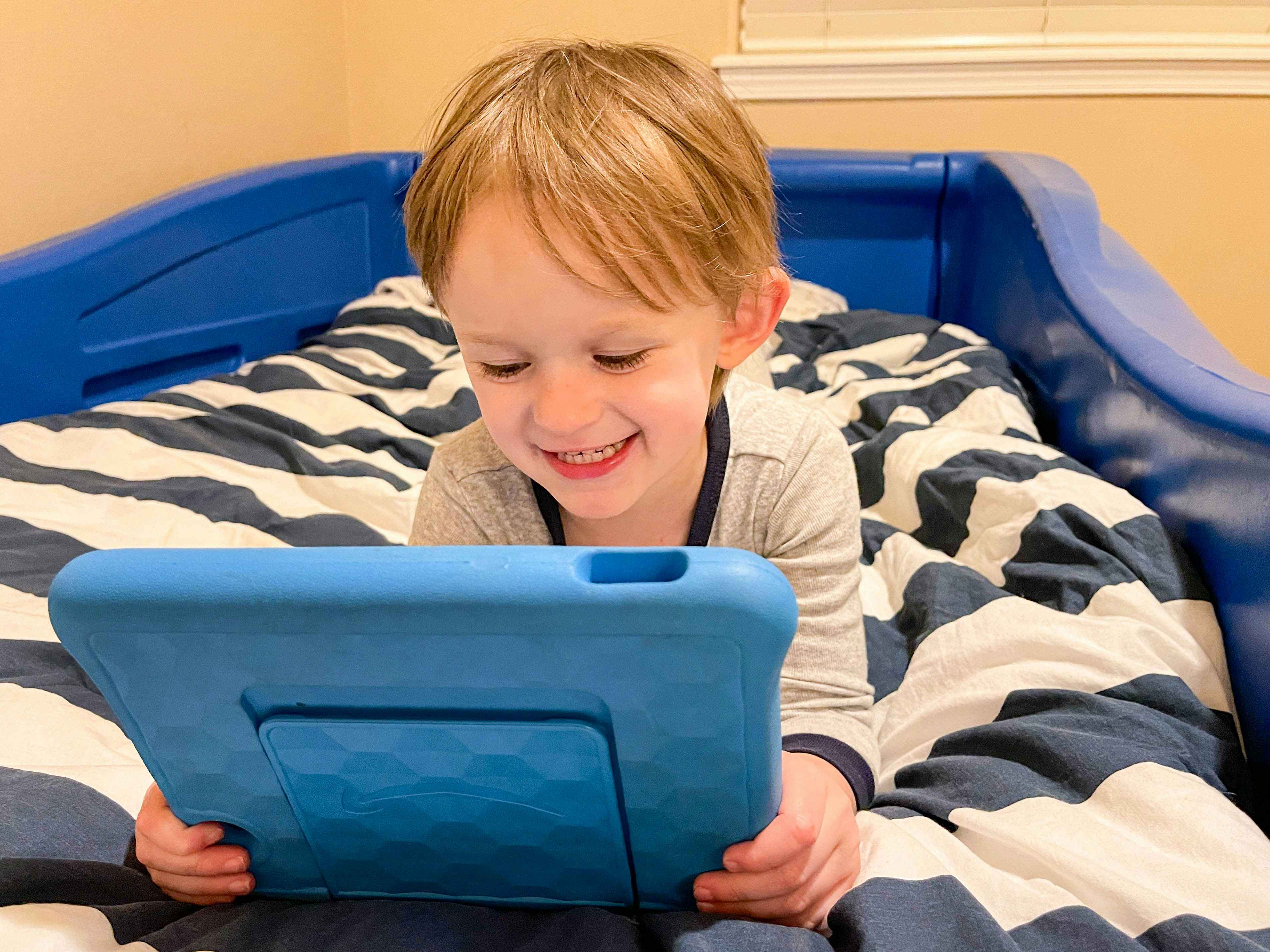 A little boy laying on a bed, watching something on an Amazon Fire tablet.