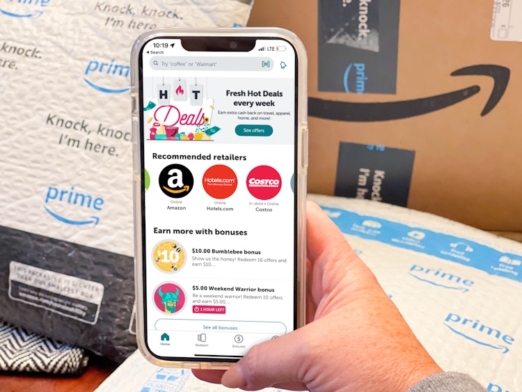 A cellphone displaying the ibotta app being held in front of Amazon boxes and packages