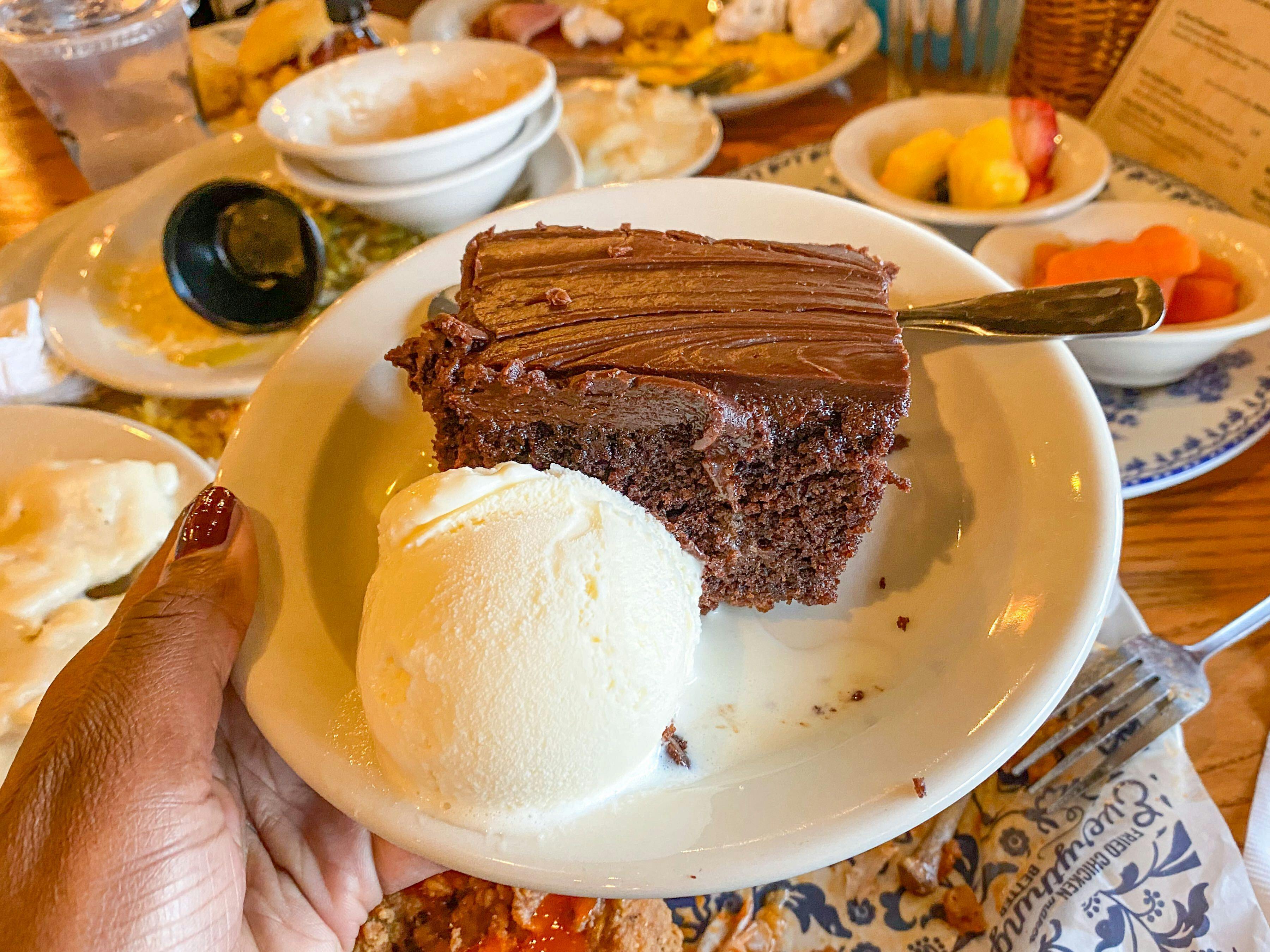 A person's hand holding up a plate with a Cracker Barrel brownie and ice cream dessert on it.