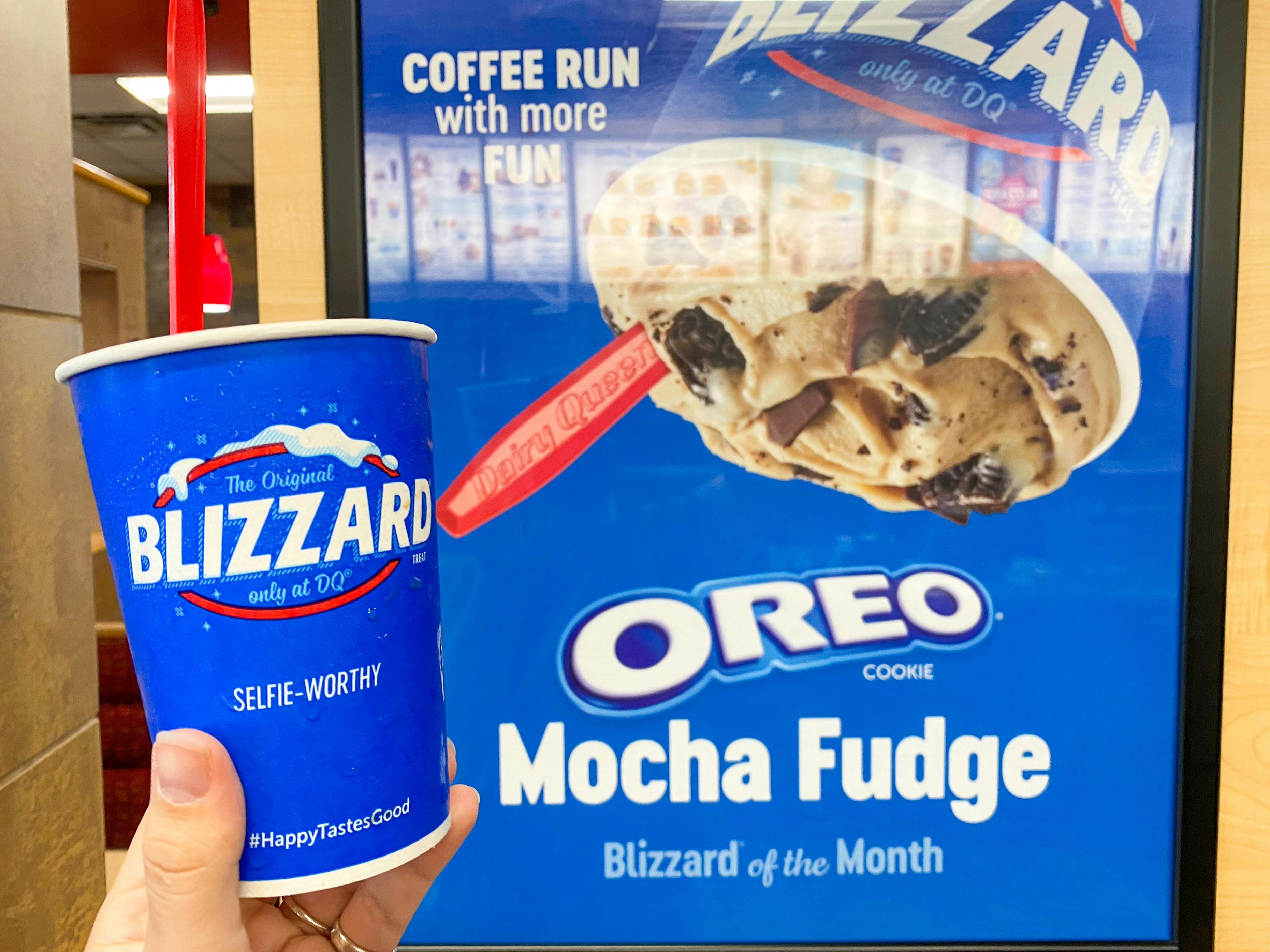 A person's hand holding up a Blizzard cup next to a poster for the Blizzard of the Month inside Dairy Queen.