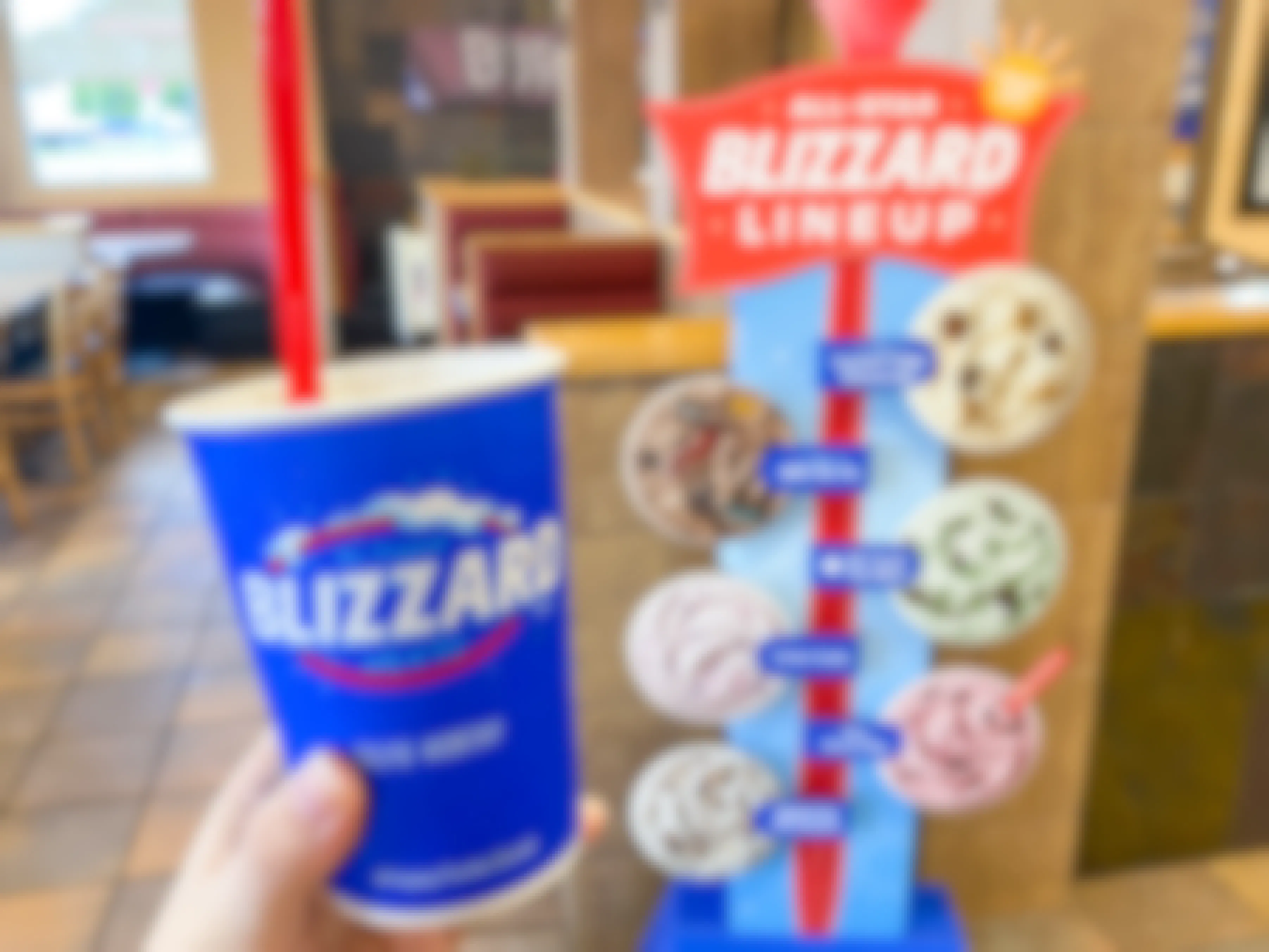 A person's hand holding up a Blizzard cup next to the All-Star Blizzard Lineup cardboard cutout displayed inside Dairy Queen.