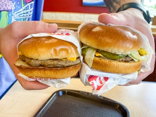 A person holding two Dairy Queen cheeseburgers next to each other above a table at Dairy Queen.