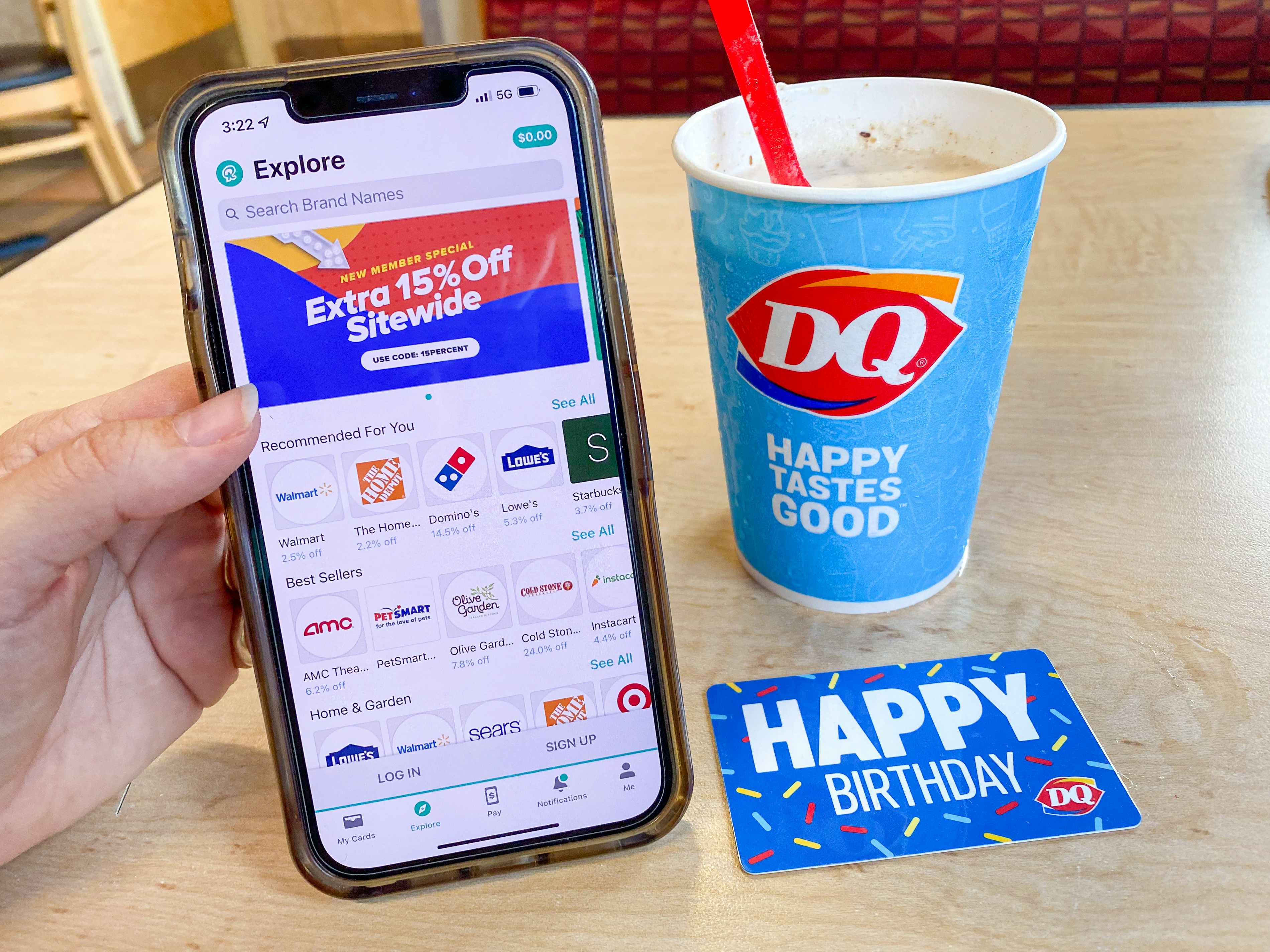 A person's hand holding a cell phone displaying the Raise.com app next to a Dairy Queen gift card and Blizzard cup sitting on a table at Dairy Queen.