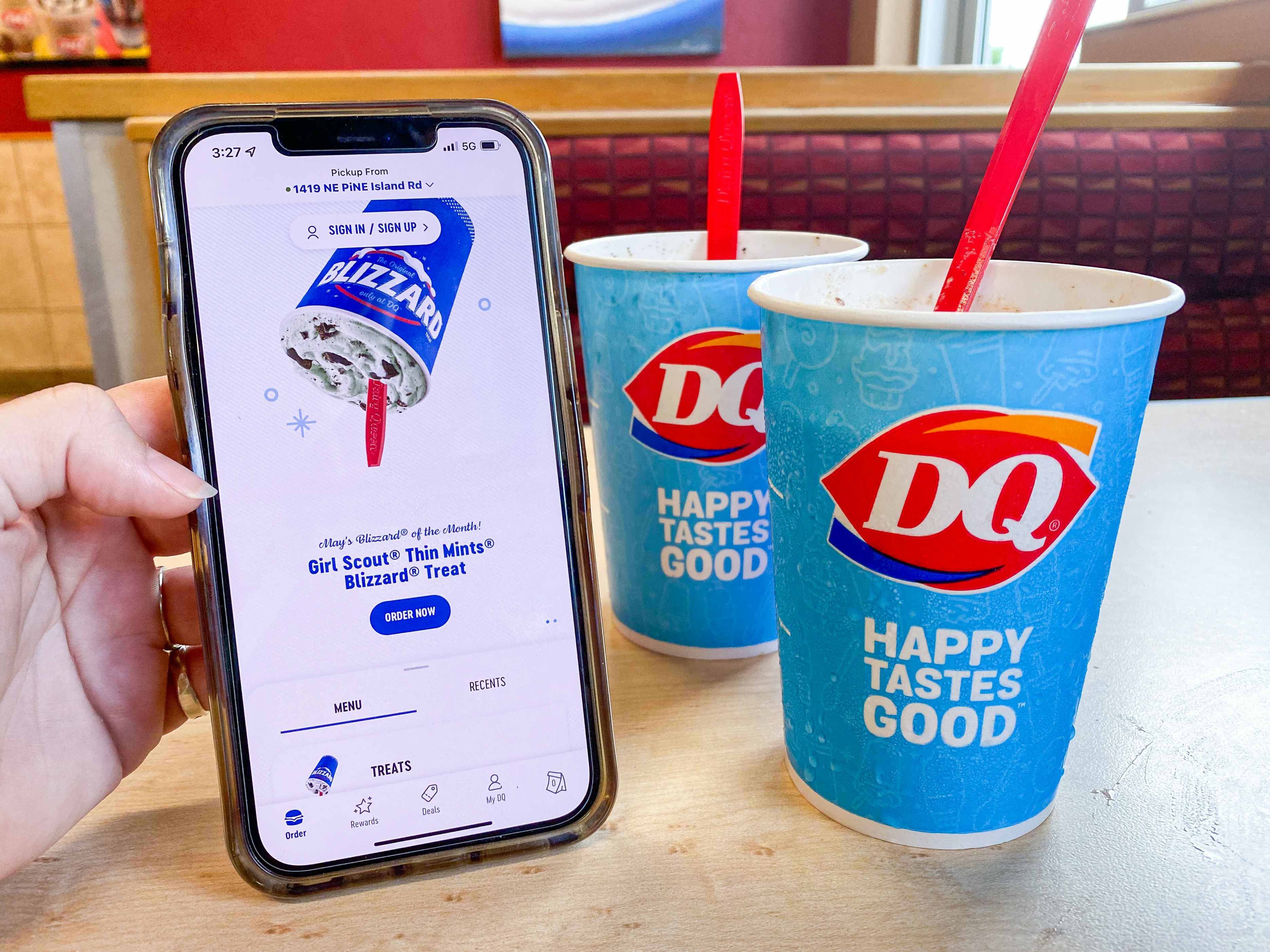 A person's hand holding a cell phone displaying the Dairy Queen app next to two Blizzard cups sitting on a table at Dairy Queen. Blizzards are some of the free fast food options available.