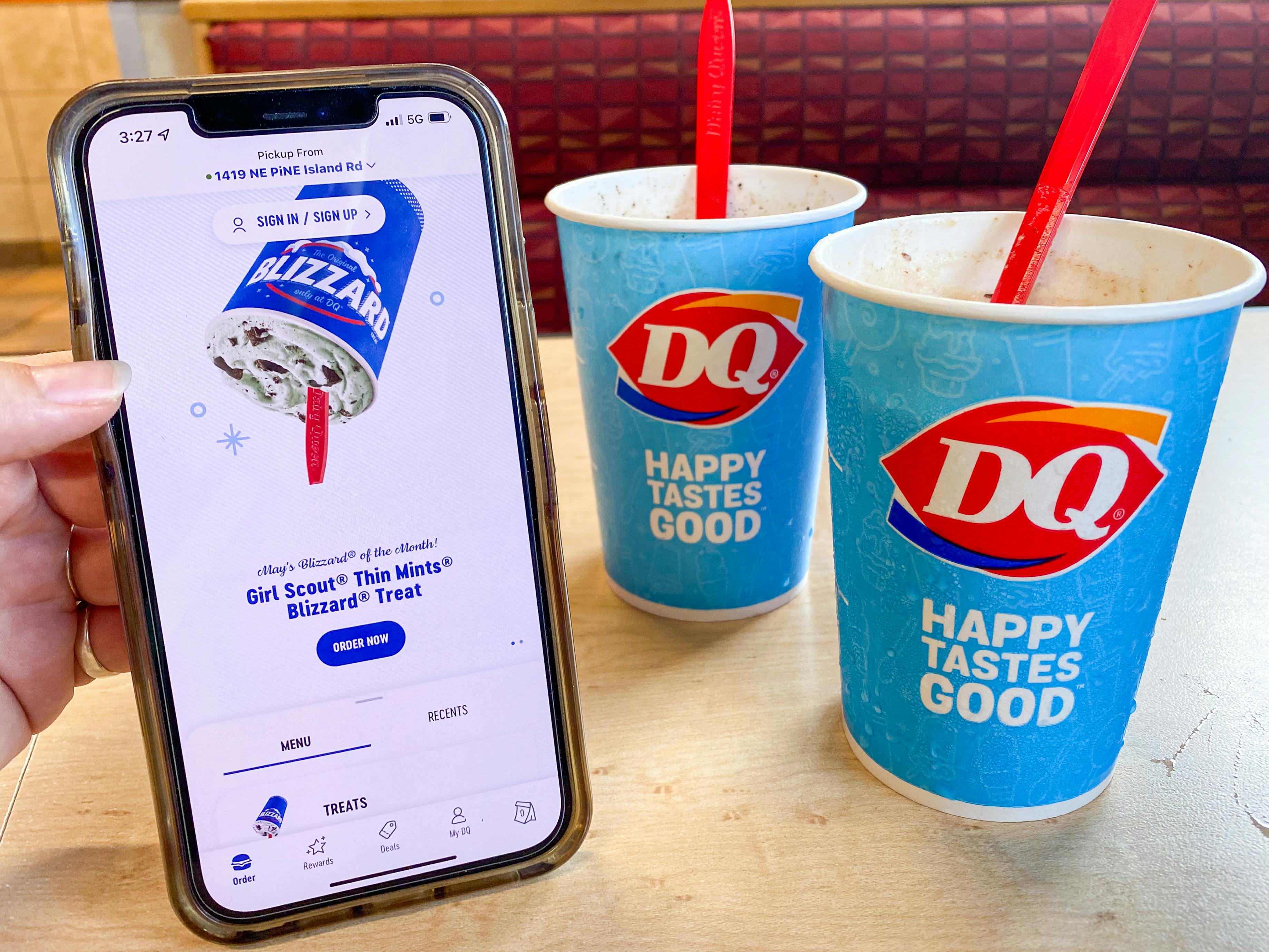 A person's hand holding a cell phone displaying the Dairy Queen app next to two Blizzard cups sitting on a table at Dairy Queen.