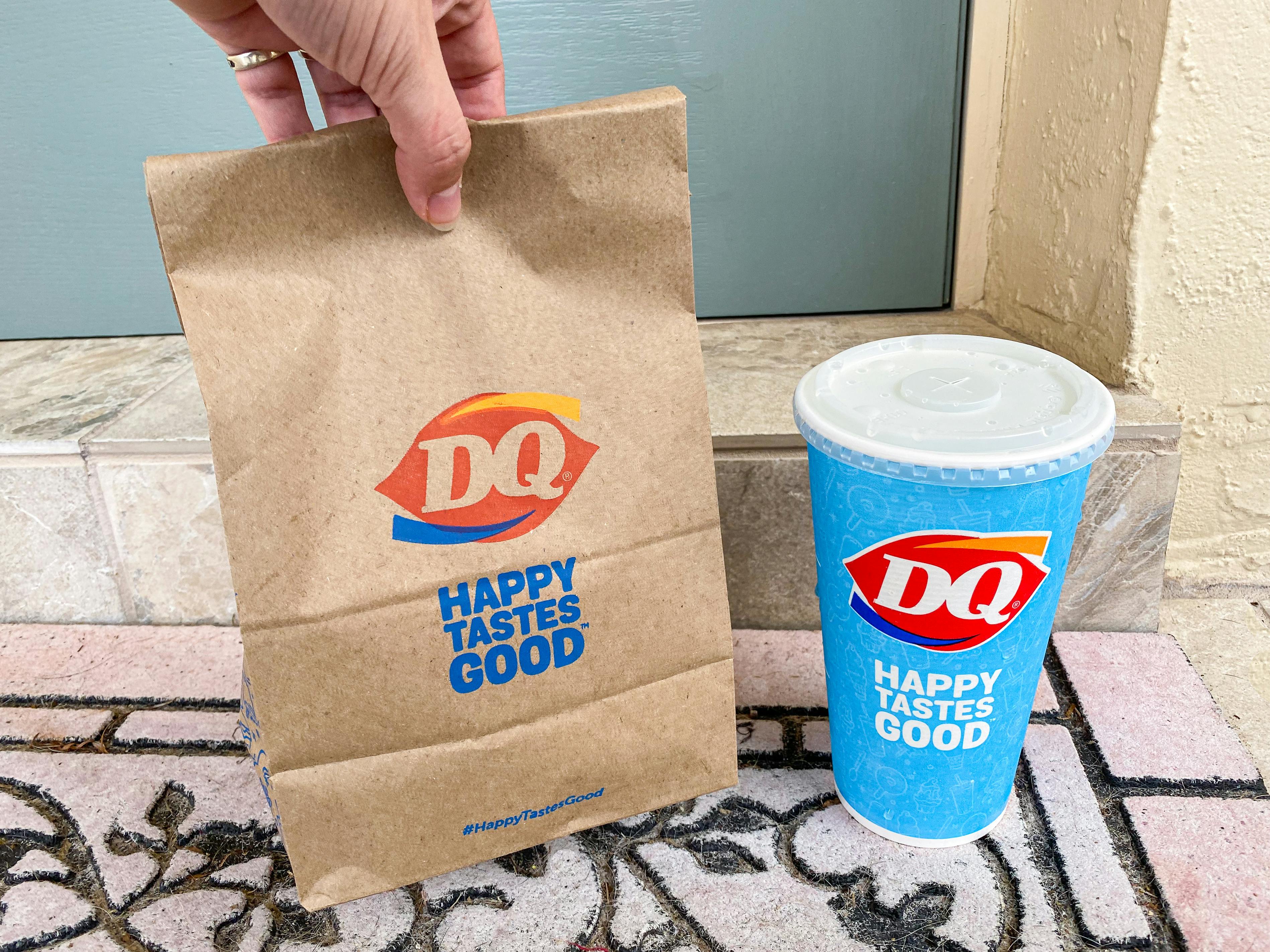 A person's hand setting a Dairy Queen takeout bag down next to a Dairy Queen drink cup sitting on a front doorstep.