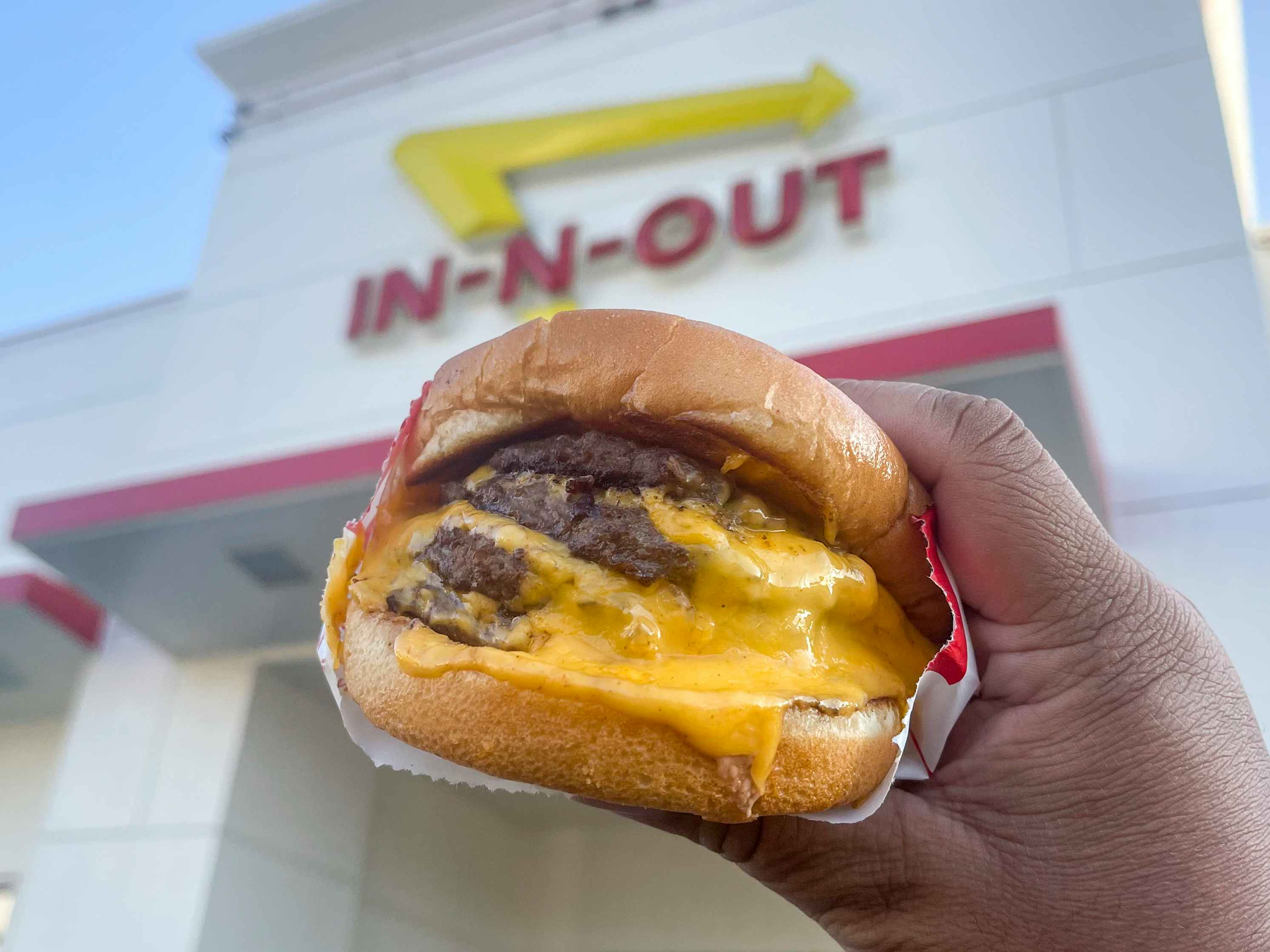 A person holding a cheeseburger with 4 patties on it in front of an In-N-Out restaurant.