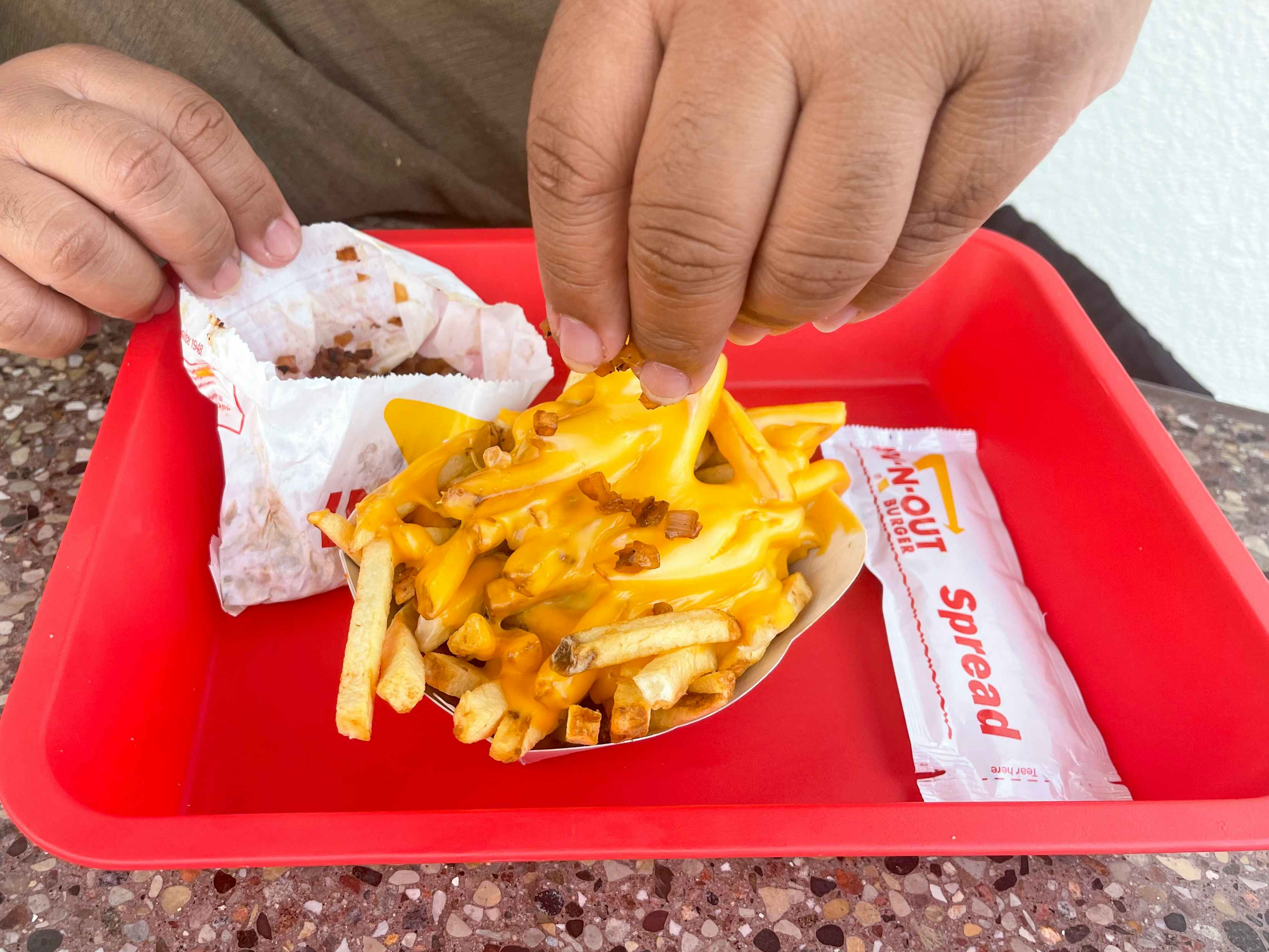 A person adding grilled onion to an order of french fries with melted cheese on top, with a packet of In-N-Out spread on the tray beside the fries, part of the in n out secret menu.