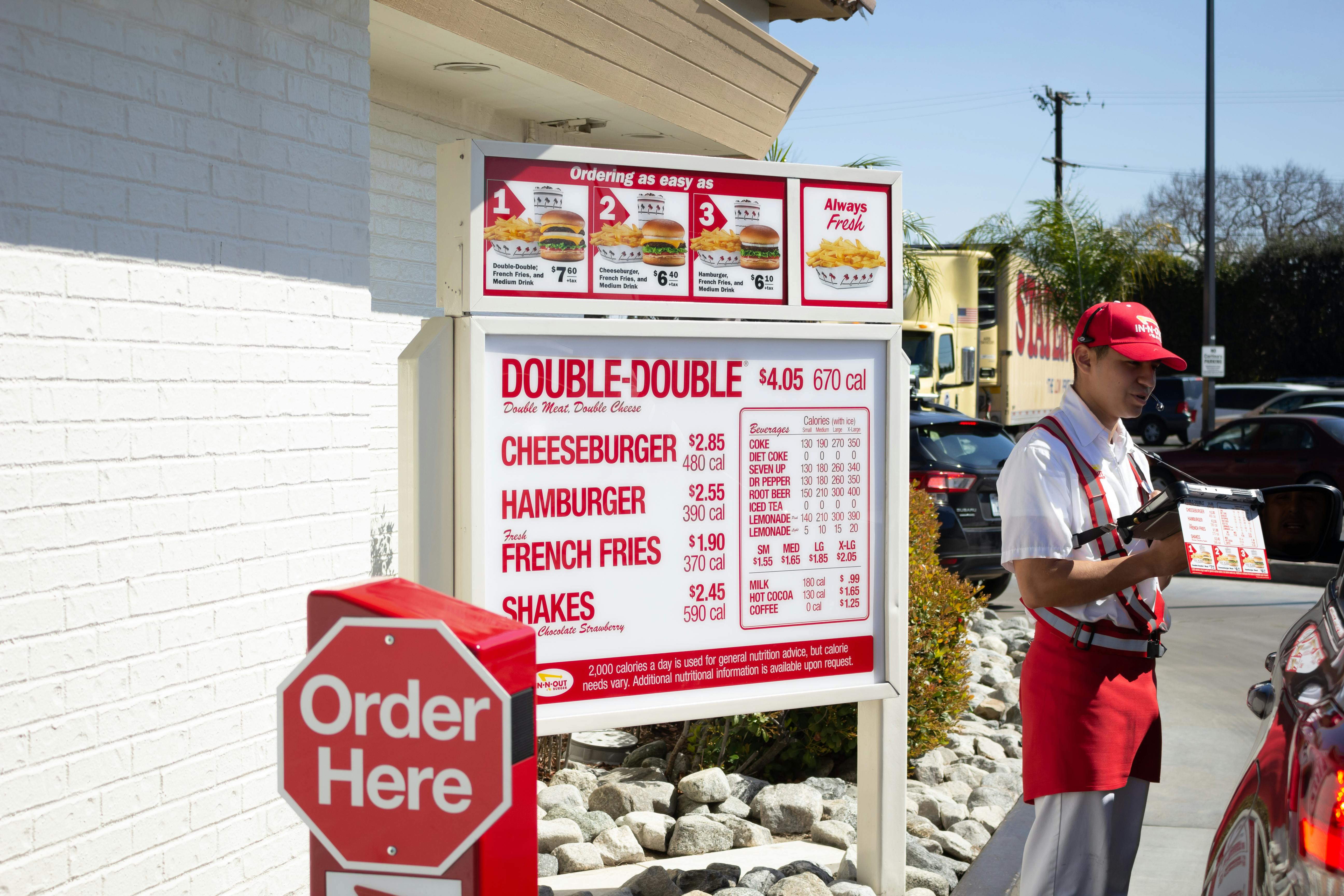 The In-N-Out drive-thru menu next to an Order Here sign and an employee talking to someone in a vehicle.