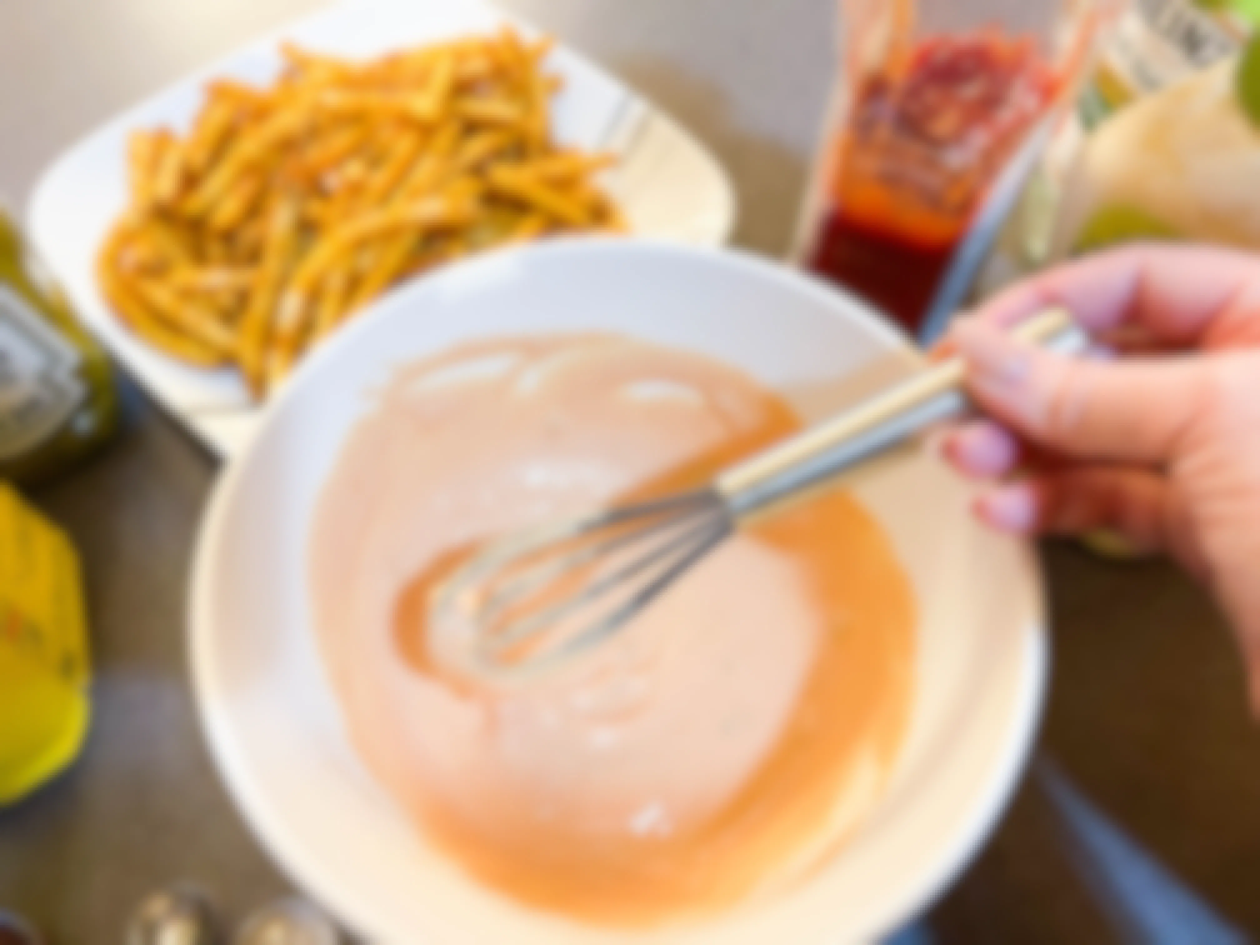 A person whisking homemade In-N-Out burger fry sauce in a mixing bowl next to a plate of fries.