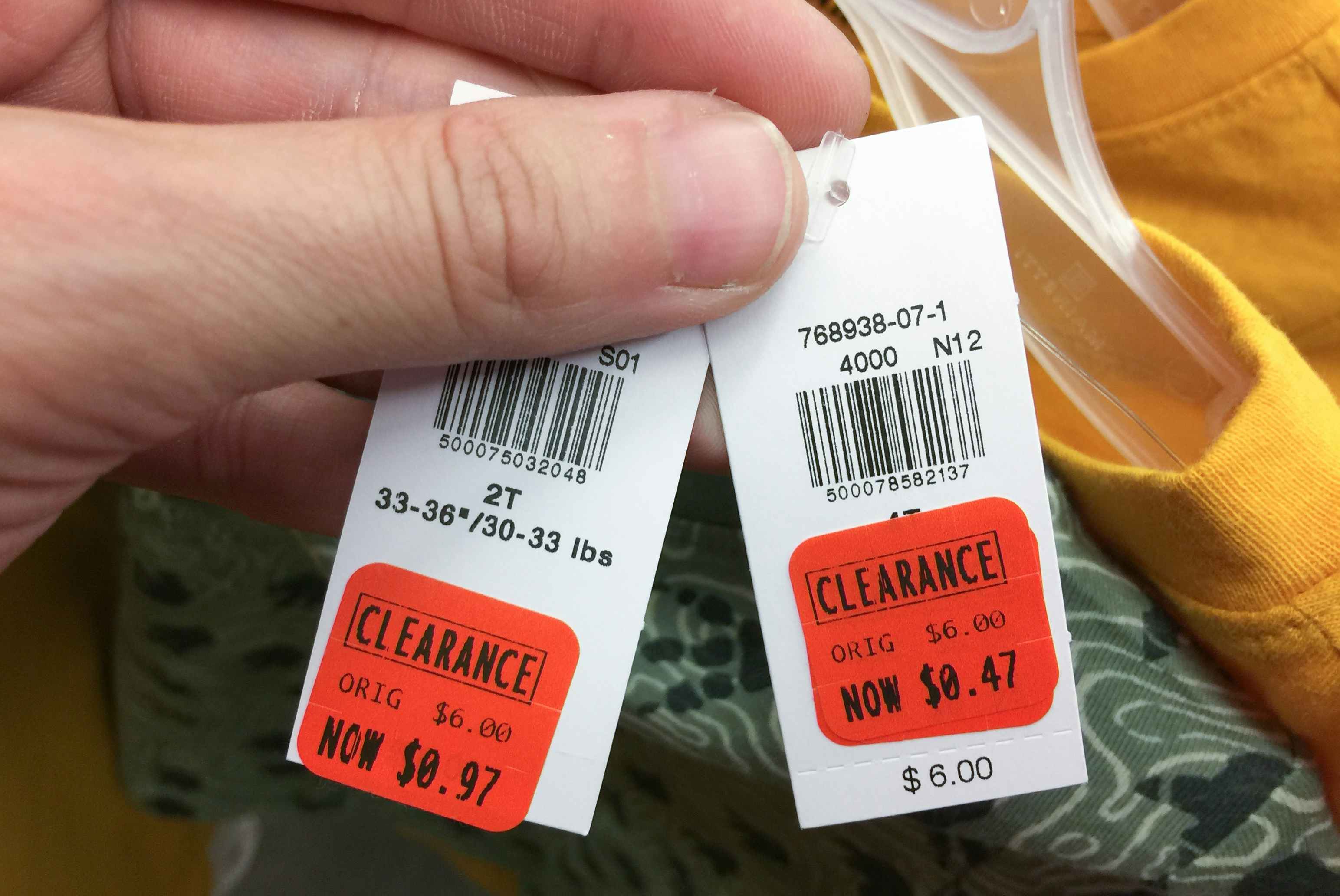 A person's hand holding up two clearance-stickered price tags, showing their marked-down prices, one being $0.97 and the other being $0.47