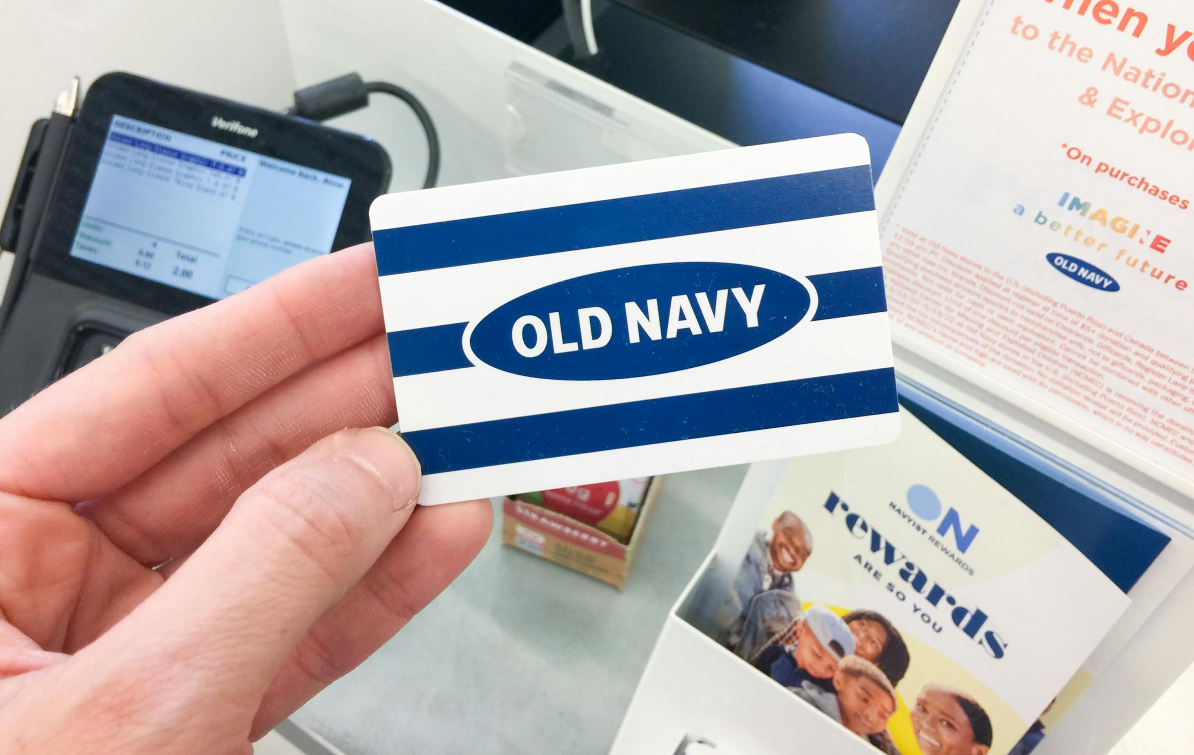 A person's hand holding an Old Navy gift card in front of the checkout counter inside Old Navy.