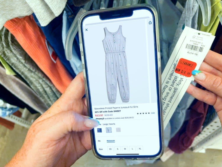 A person's hand holding a cell phone displaying a clothing item next to a tag of the same clothing item in-store at Old Navy.