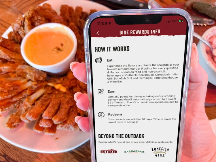 A person's hand holding their phone displaying Outback Steakhouse's Dine Rewards Info in front of a plate of a Bloomin' Onion 