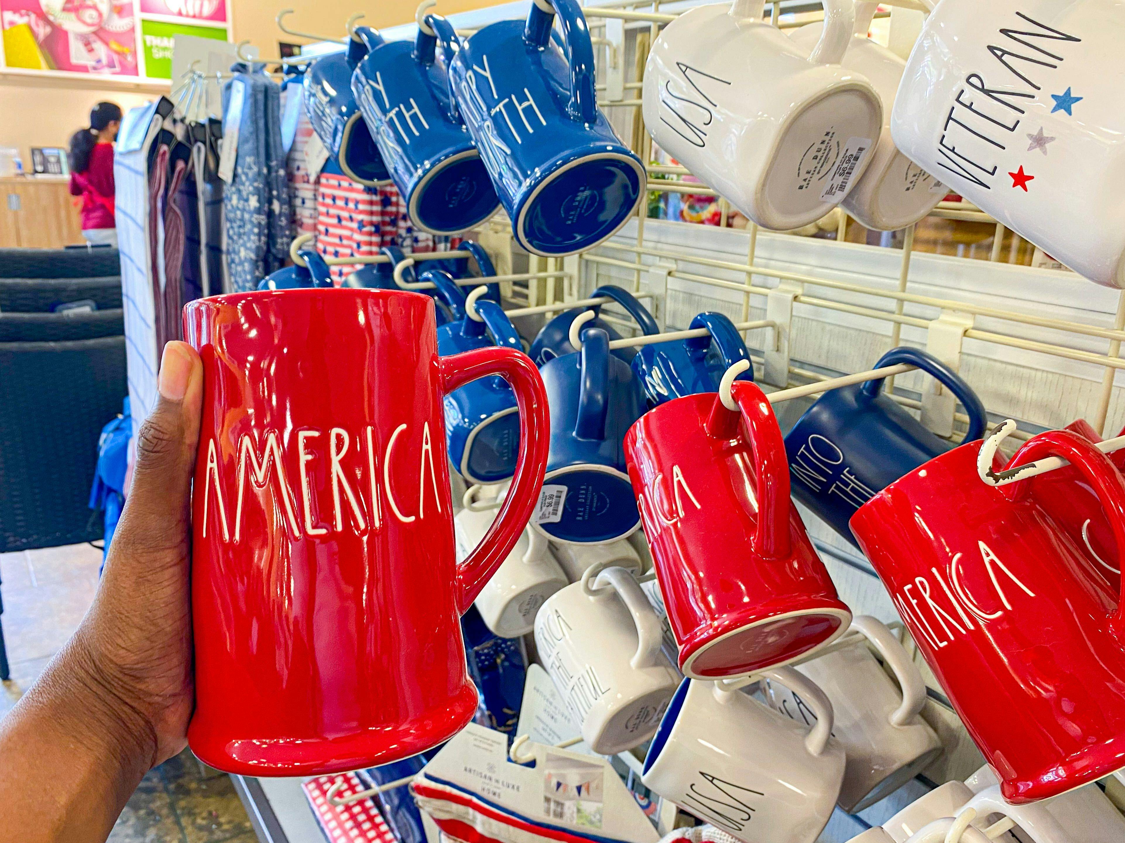 A person's hand holding a Rae Dunn mug that says "America" in front of a display of Americana Rae Dunn mugs.