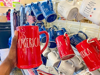 A person's hand holding a Rae Dunn mug that says "America" in front of a display of Americana Rae Dunn mugs.