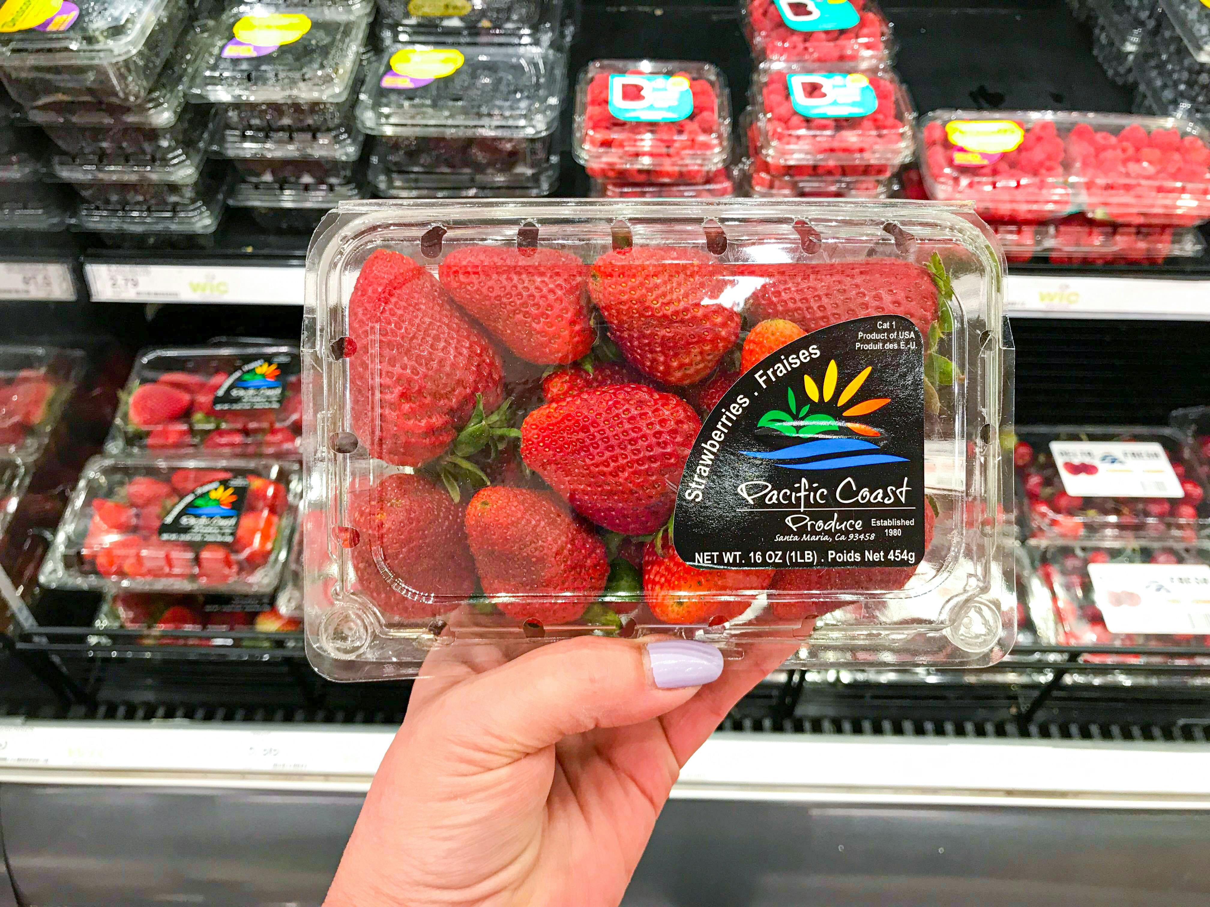 Someone holding a package of strawberries in front of a refrigerated shelf of more fruit in a grocery store.