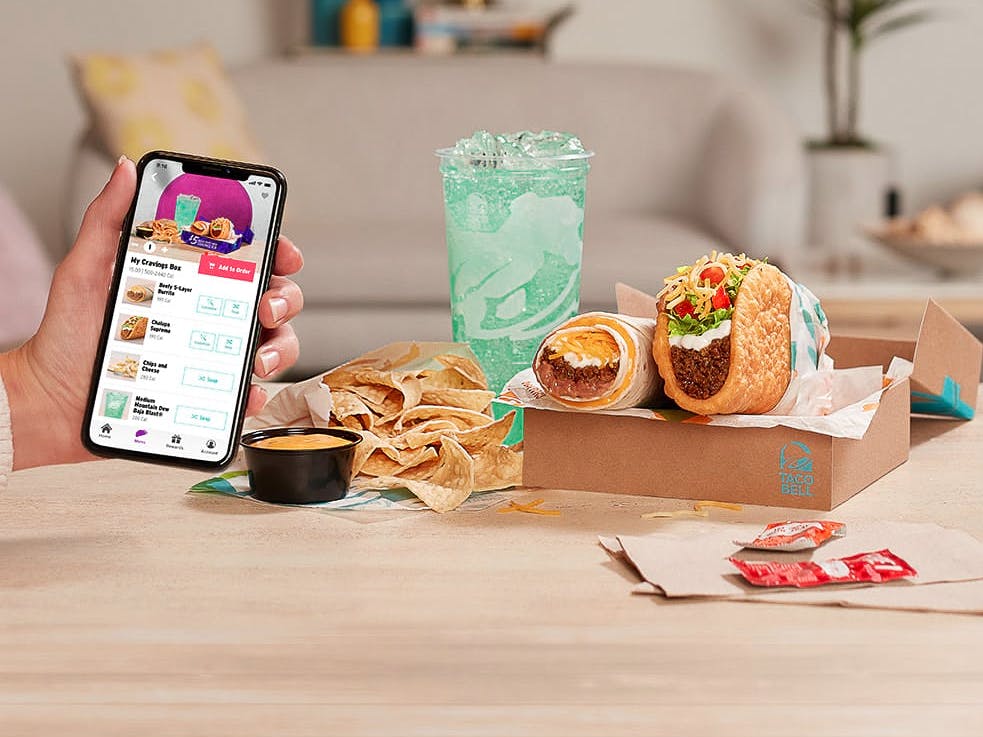 A person's hand holding up their phone displaying the Build your Own Cravings Box menu on the Taco Bell app next to food from a Cravings Box sitting on a table.