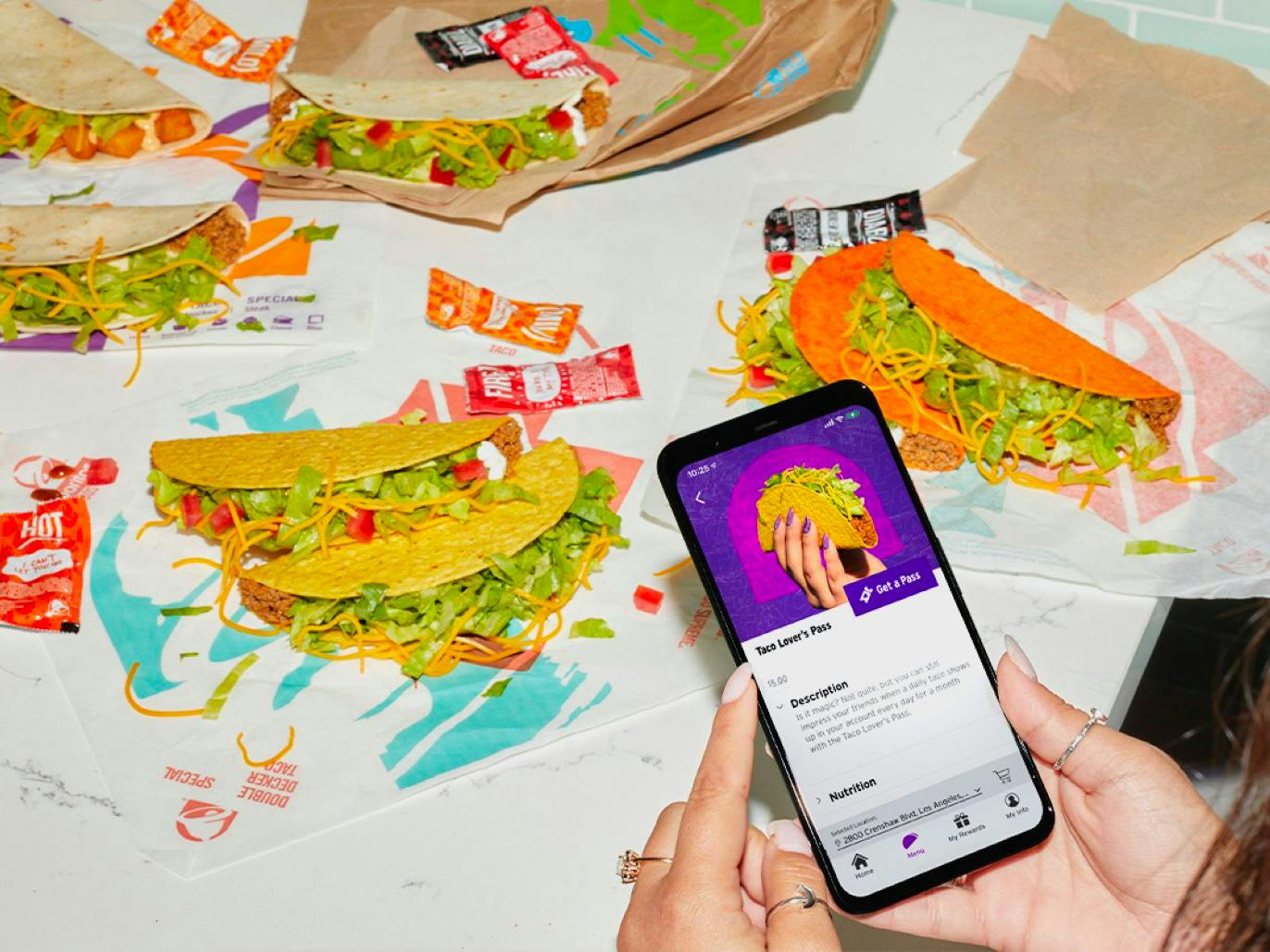A person's hands holding their phone displaying the Taco Lovers Pass on the Taco Bell app in front of a table of unwrapped tacos.