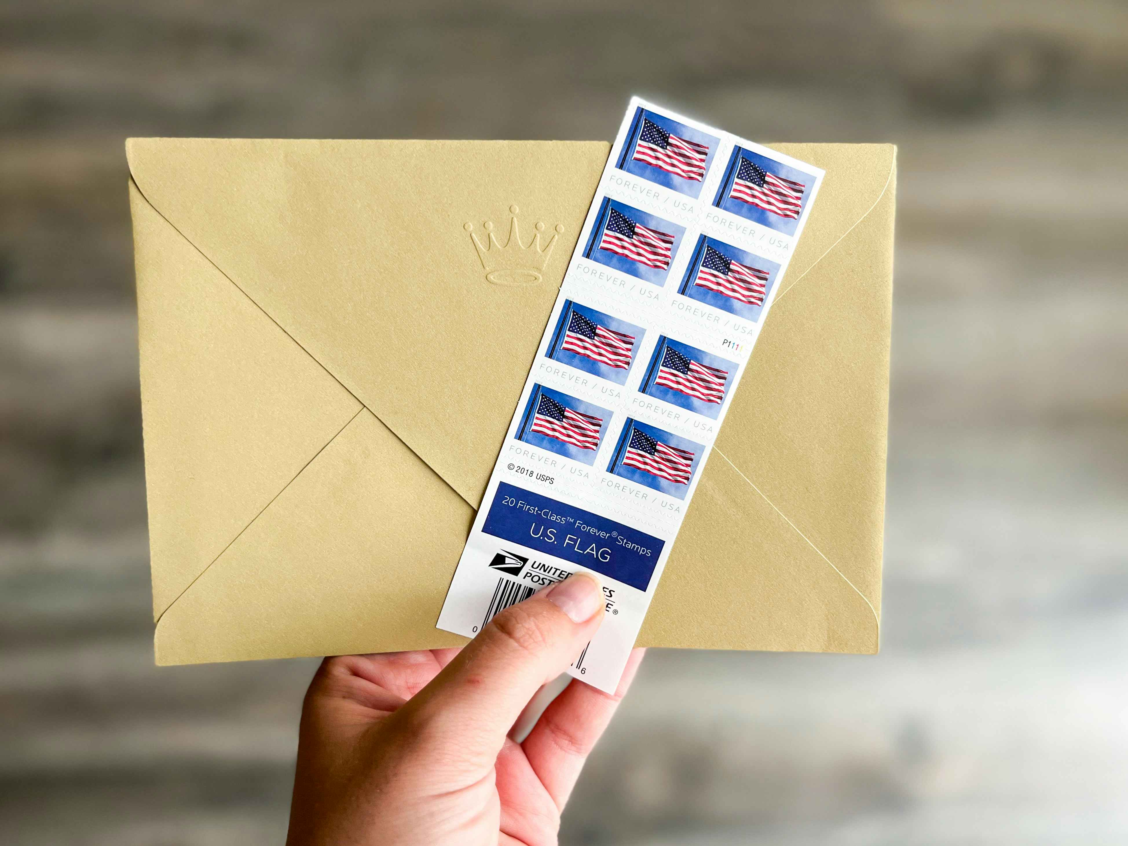 A person's hand holding up an envelope and some American flag Forever postage stamps.