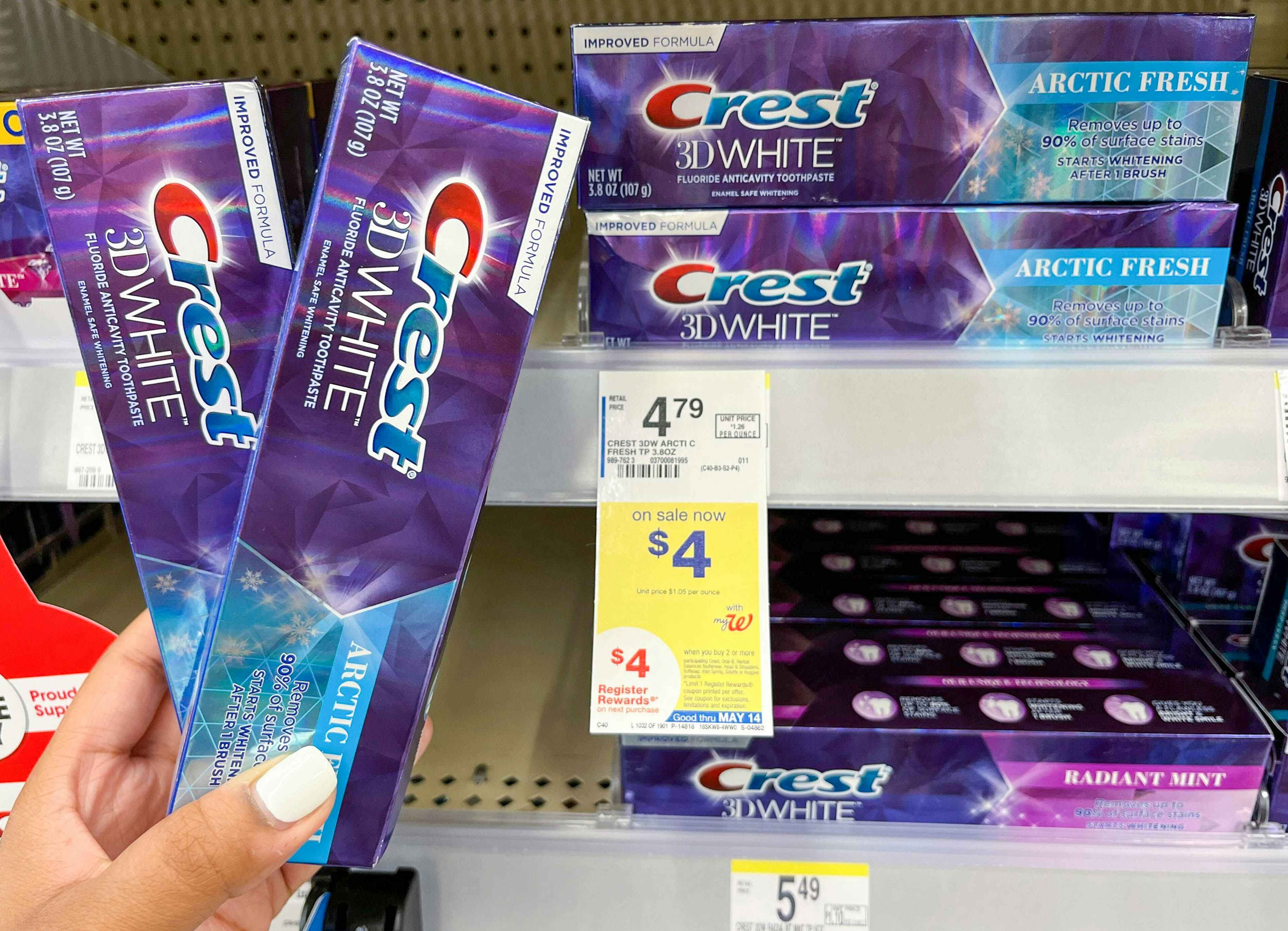 A person's hand holding two boxes of Crest toothpaste next to a sale tag at Walgreens.