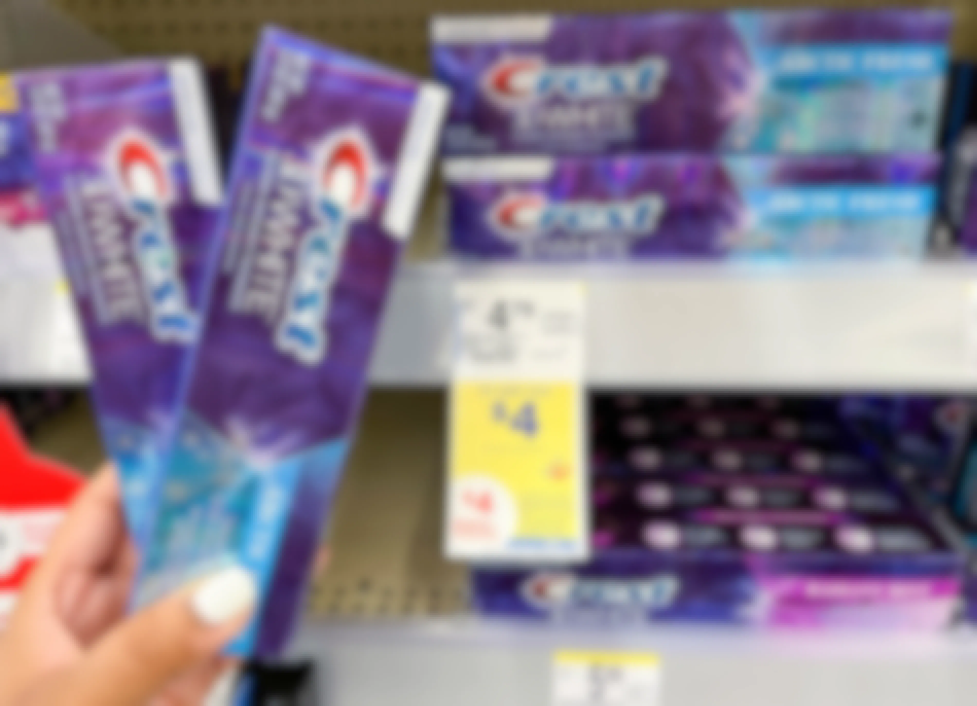 A person's hand holding two boxes of Crest toothpaste next to a sale tag at Walgreens.