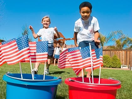 Two kids in the backyard running toward a red and blue bucket of American flags.