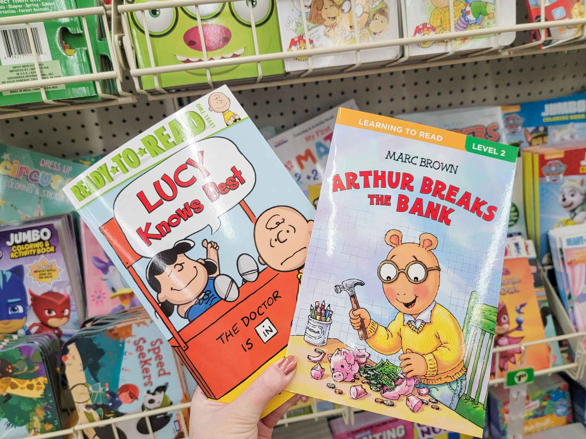 hand holding two kids books: lucy knows best and arthur breaks the bank