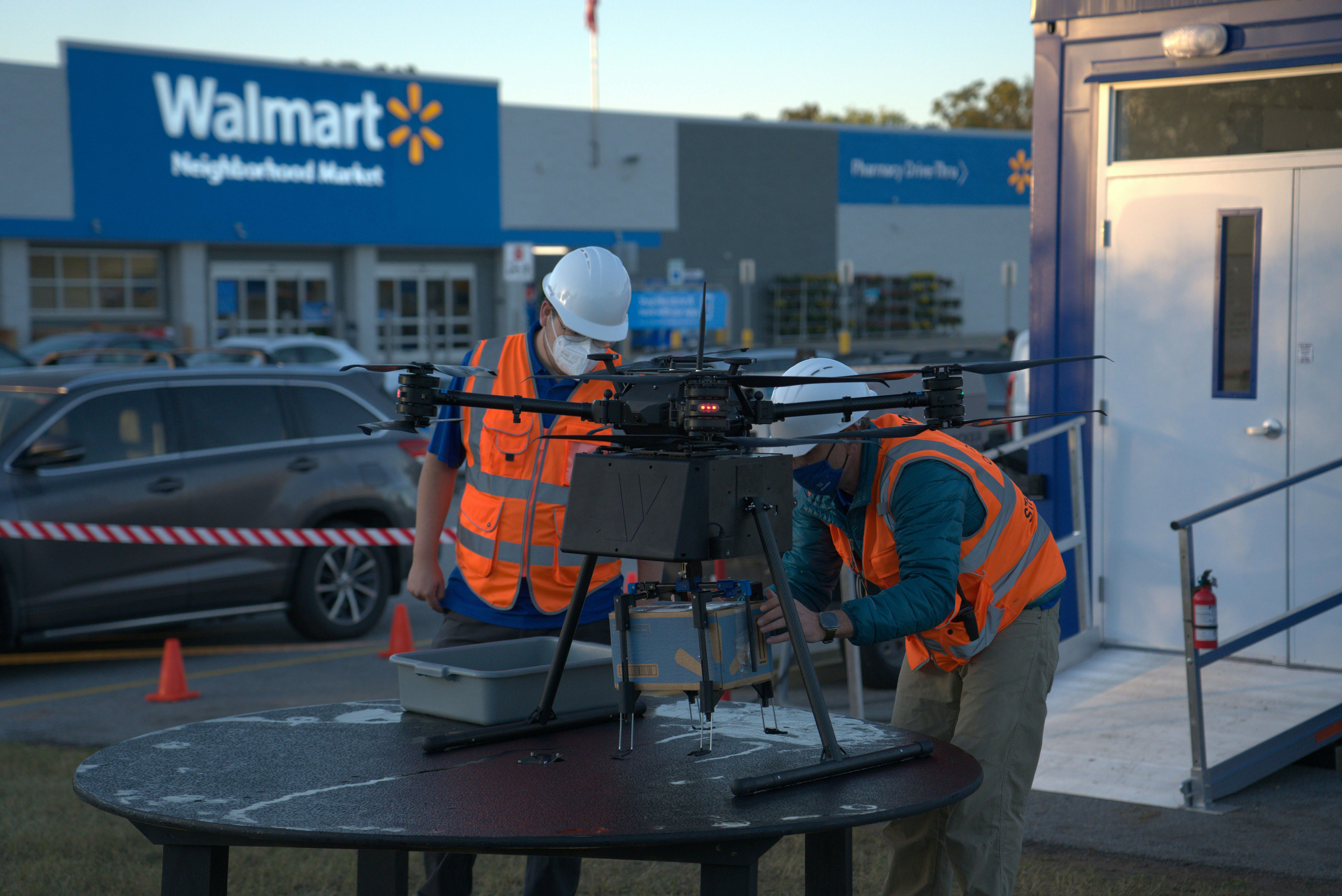 Two people in visibility vests attaching a Walmart delivery box to a drone that is parked on a table in the parking lot outside Walmart.
